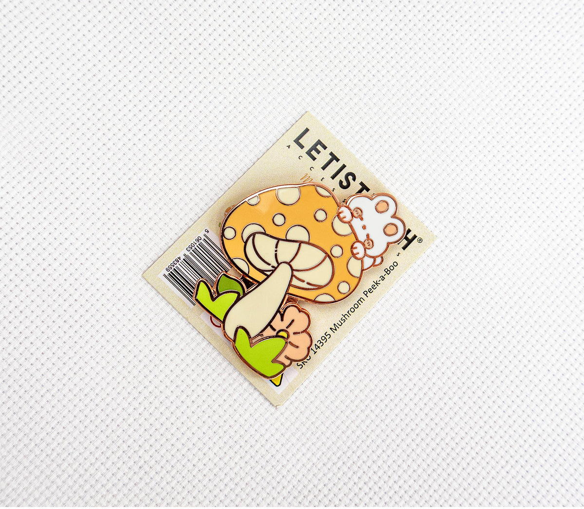 A small enamel pin shaped like a smiling orange mushroom with white spots, cream stem, and green leaves, attached to a printed backing card. 