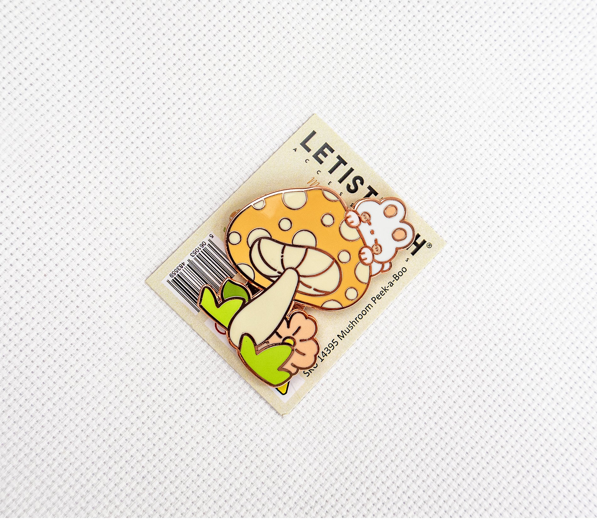 A small enamel pin shaped like a smiling orange mushroom with white spots, cream stem, and green leaves, attached to a printed backing card. 