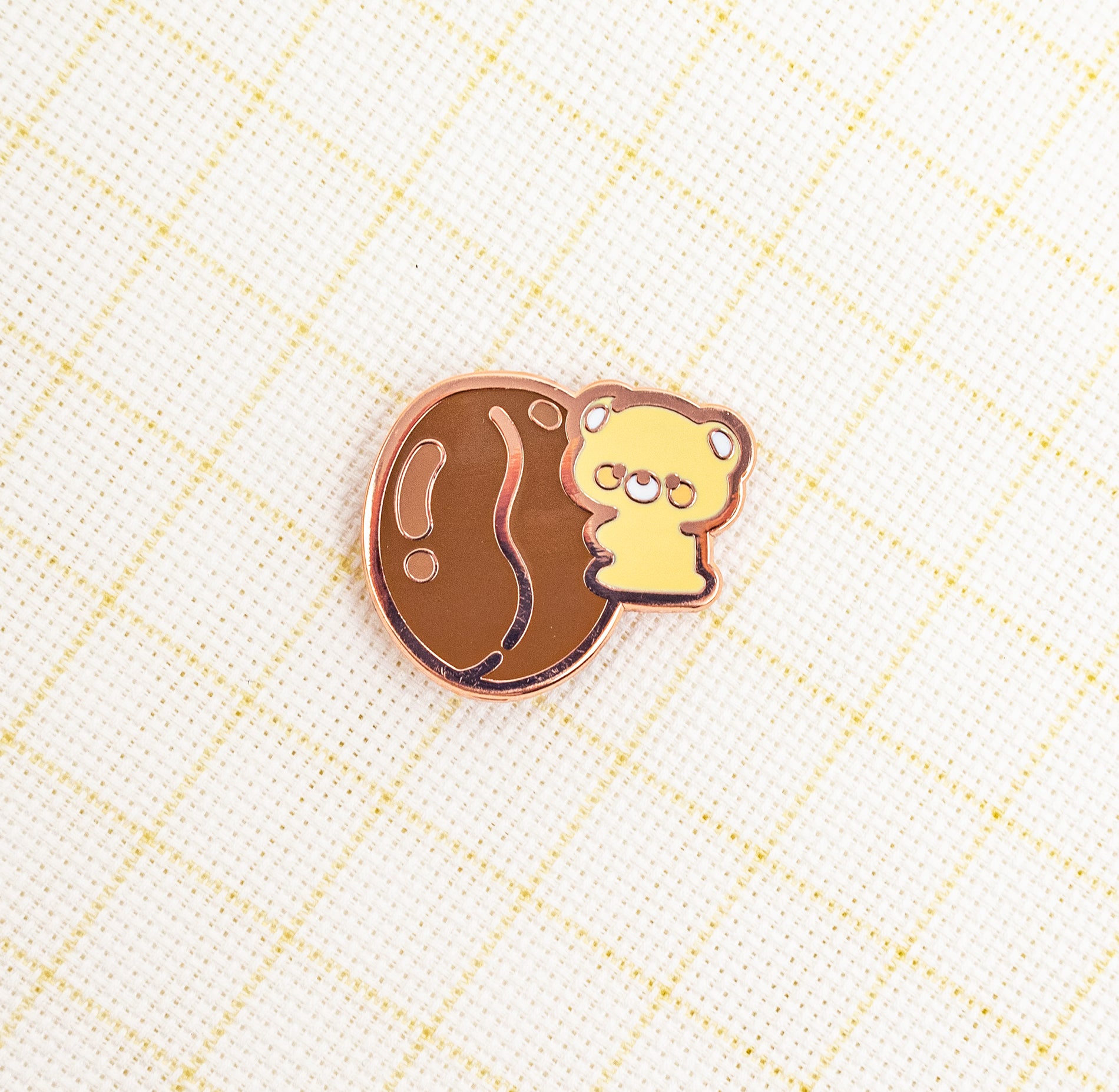 A small enamel pin featuring a yellow cartoon bear peeking out from a brown oval shape, outlined in rose gold, placed on a white textured fabric with a light yellow grid pattern.