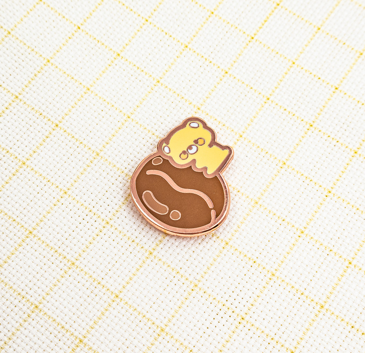 A small enamel pin featuring a yellow cartoon character peeking out of a brown cracked shell, placed on a white textured fabric with a light yellow grid pattern.