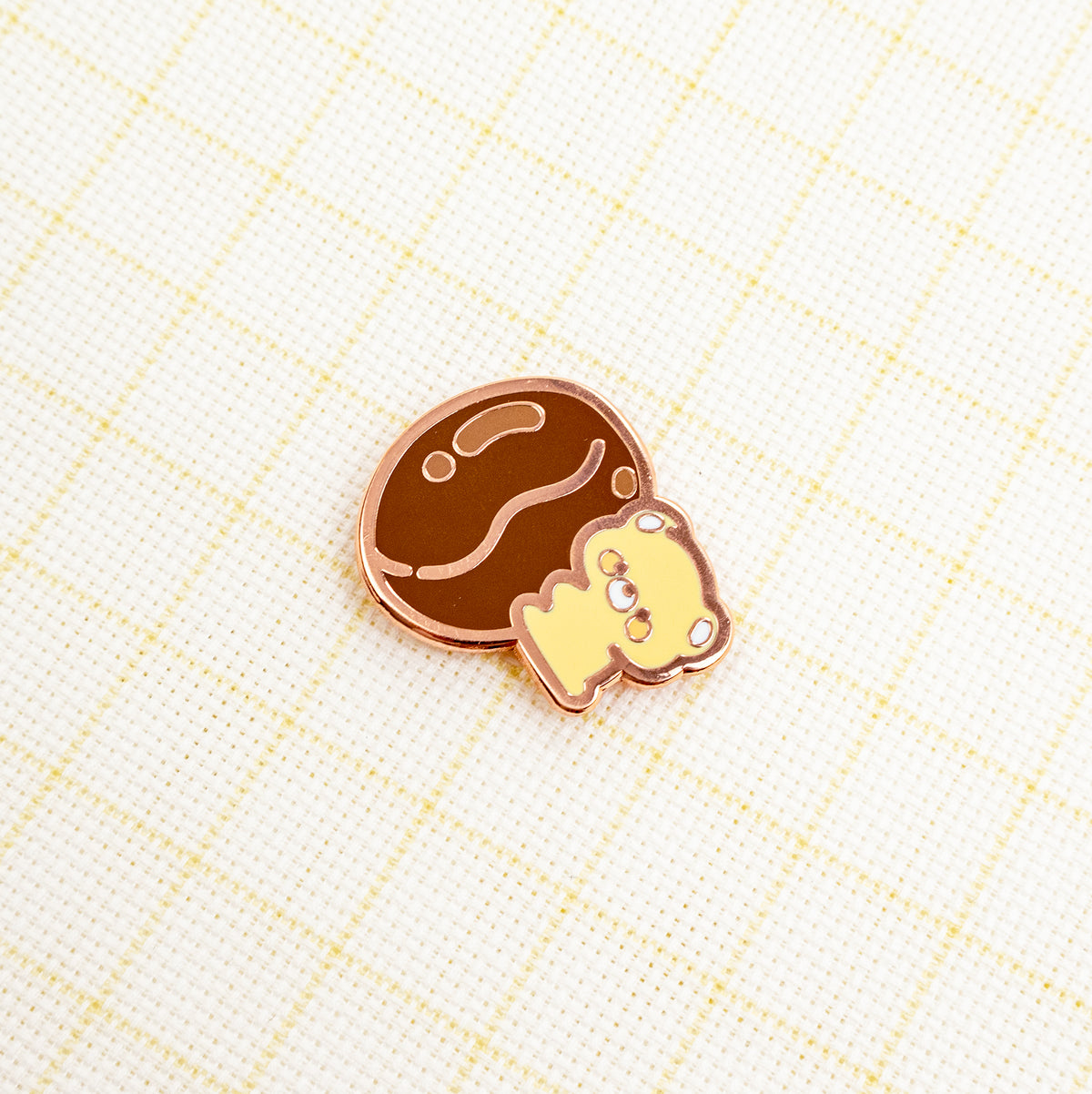 A cute enamel pin featuring a brown chocolate cookie and a small yellow bear, both outlined in rose gold metal, resting on a cream-colored textured fabric with a light yellow grid pattern.