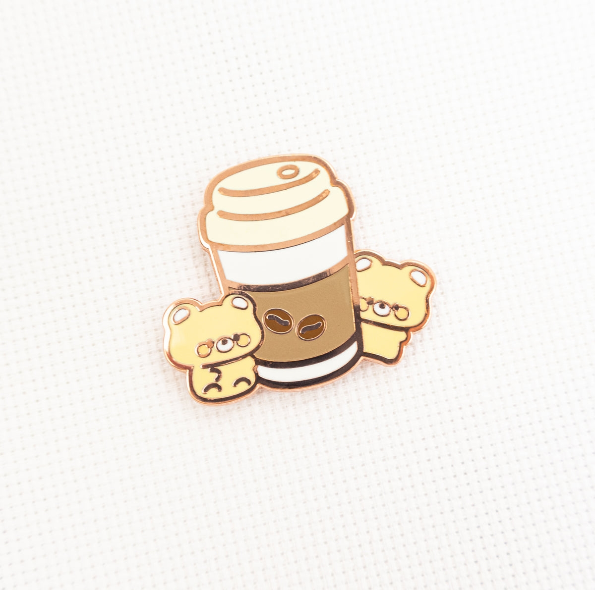 A cute enamel pin featuring a coffee cup with two small teddy bears peeking out from behind, on a textured white background.
