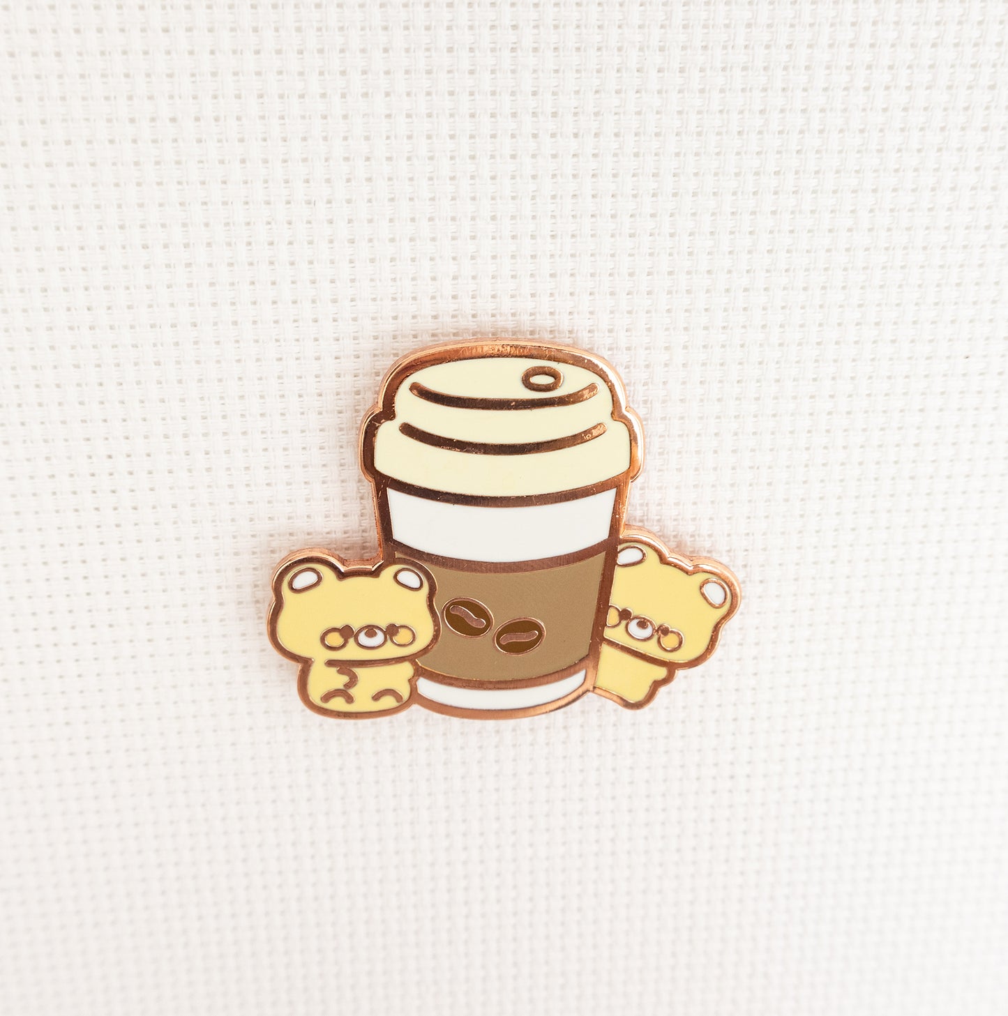 A cute enamel pin featuring a coffee cup with two small yellow teddy bears peeking out from the sides, set against a textured white background.