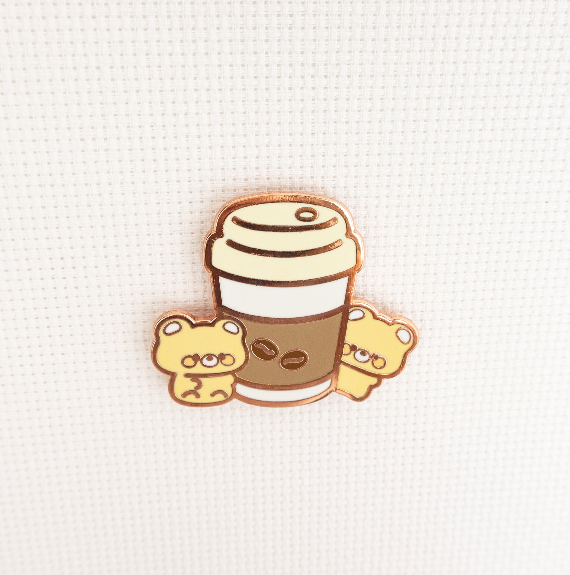 A cute enamel pin featuring a coffee cup with two small yellow teddy bears peeking out from the sides, set against a textured white background.