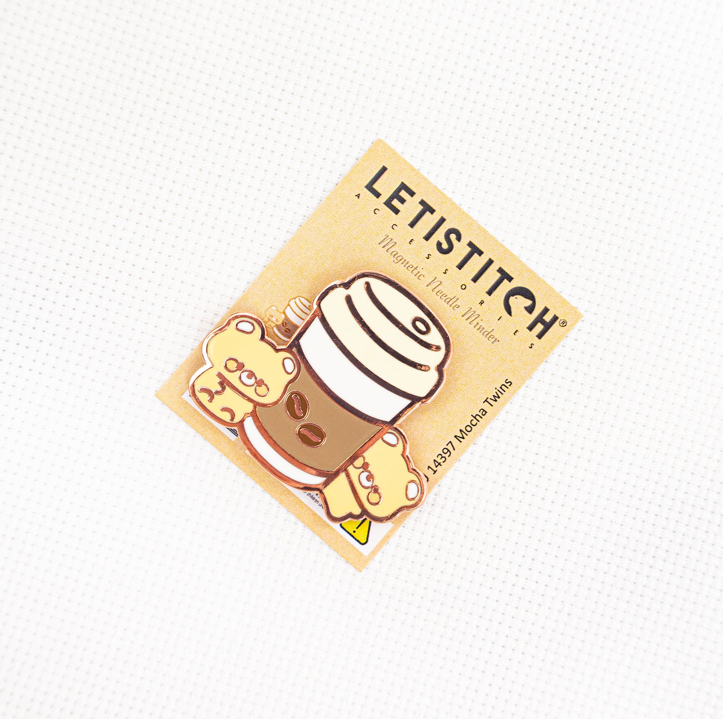 A small rectangular sticker or card featuring a cute cartoon mocha cup with a striped hat and a small character, placed on a white textured background. The card reads LETISTITCH and shows a warm beige background.
