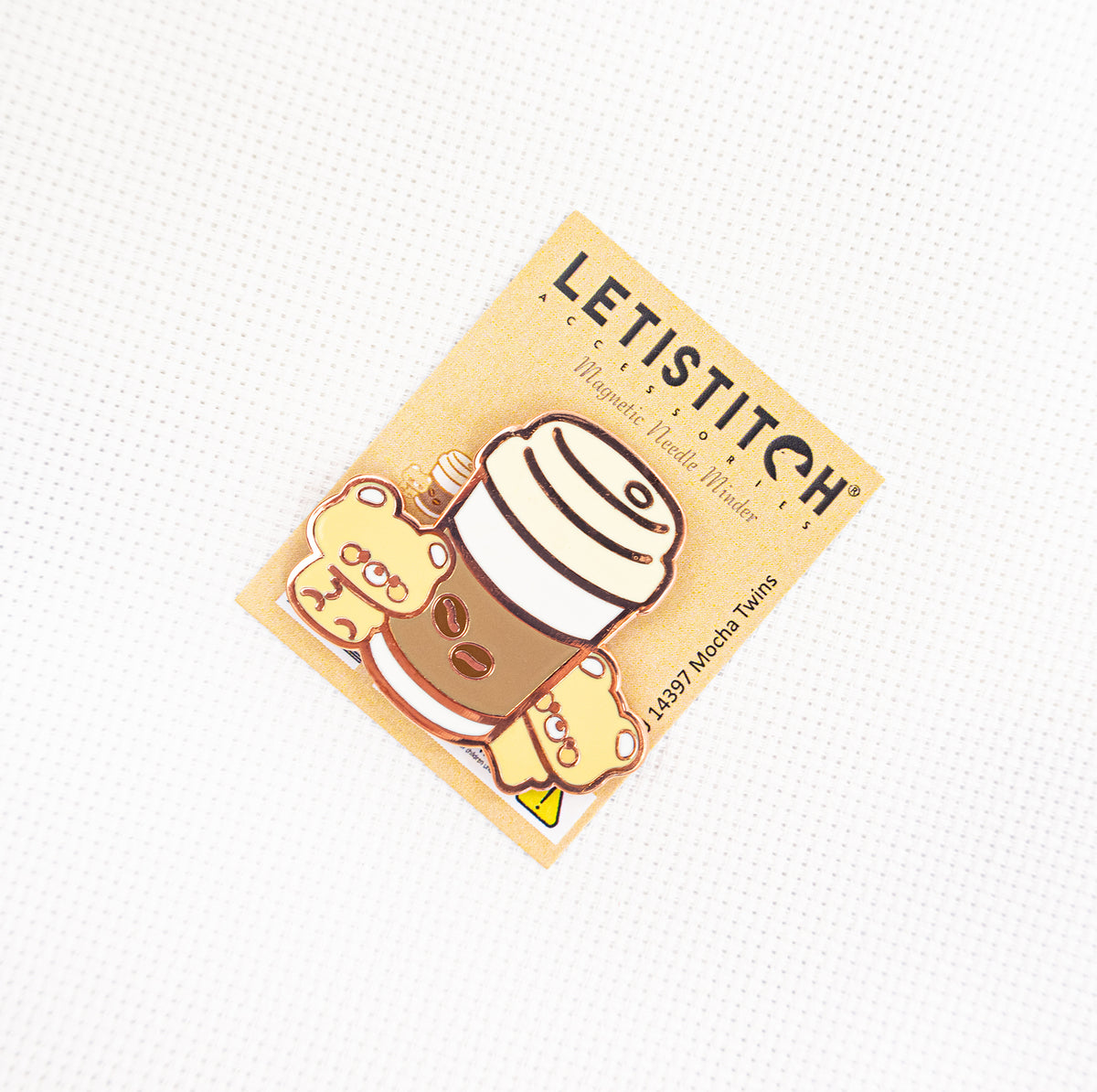 A small rectangular sticker or card featuring a cute cartoon mocha cup with a striped hat and a small character, placed on a white textured background. The card reads LETISTITCH and shows a warm beige background.