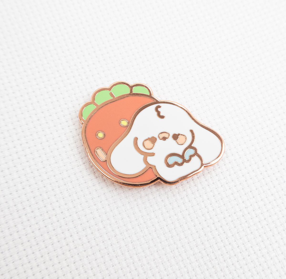 A cute enamel pin featuring a white cartoon dog peeking out from behind a strawberry, with a rose gold metal border, resting on a textured white fabric background.