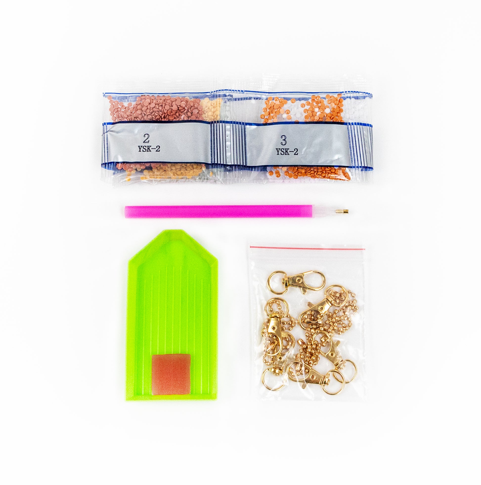 Assorted bags with crystals for diamond art including a green tag, pink tool, and gold chain on a white background.