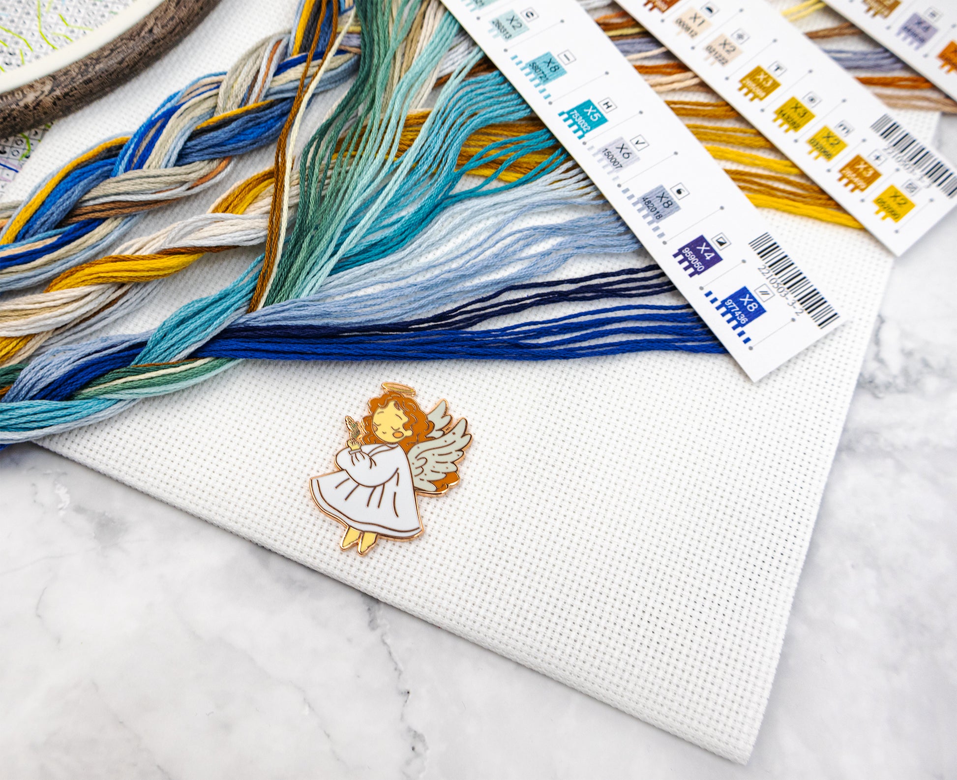 Embroidery floss with colorful strands and a small angel pin on a marble surface