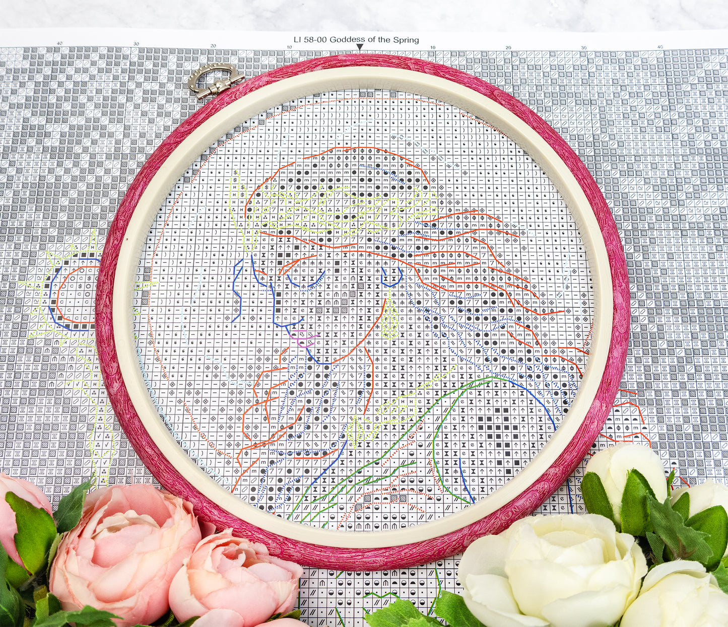 Embroidery hoop with cross-stitched design on fabric, surrounded by flowers.