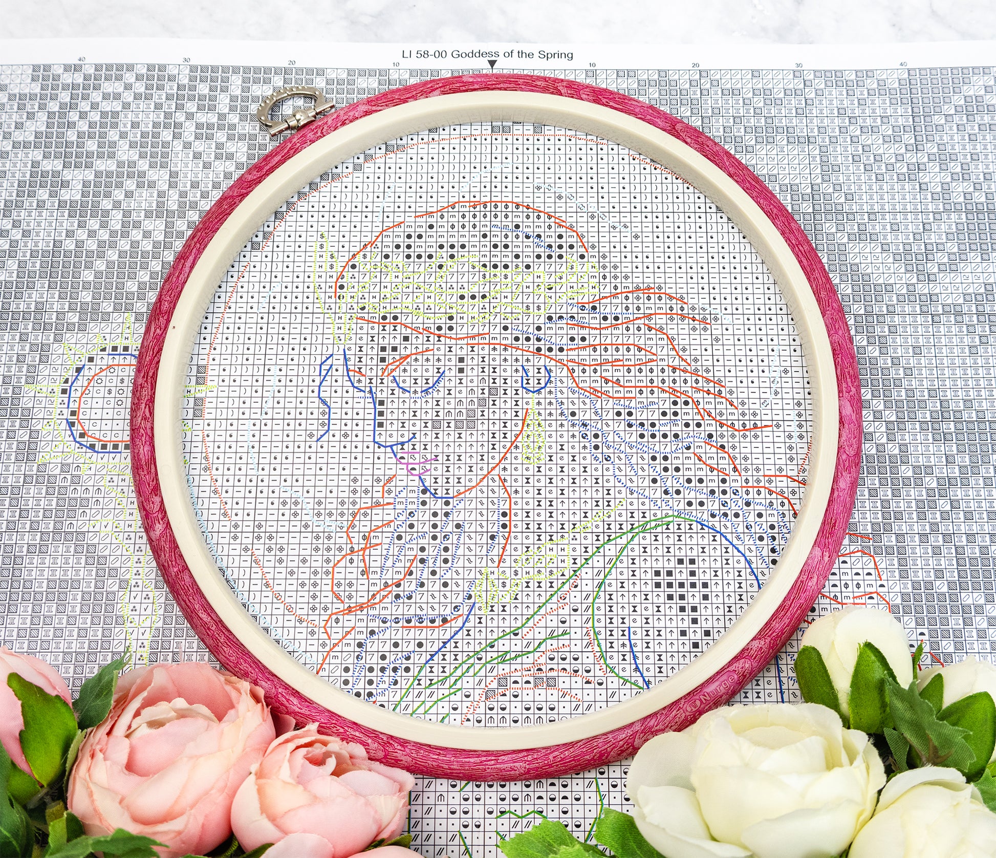 Embroidery hoop with cross-stitched design on fabric, surrounded by flowers.