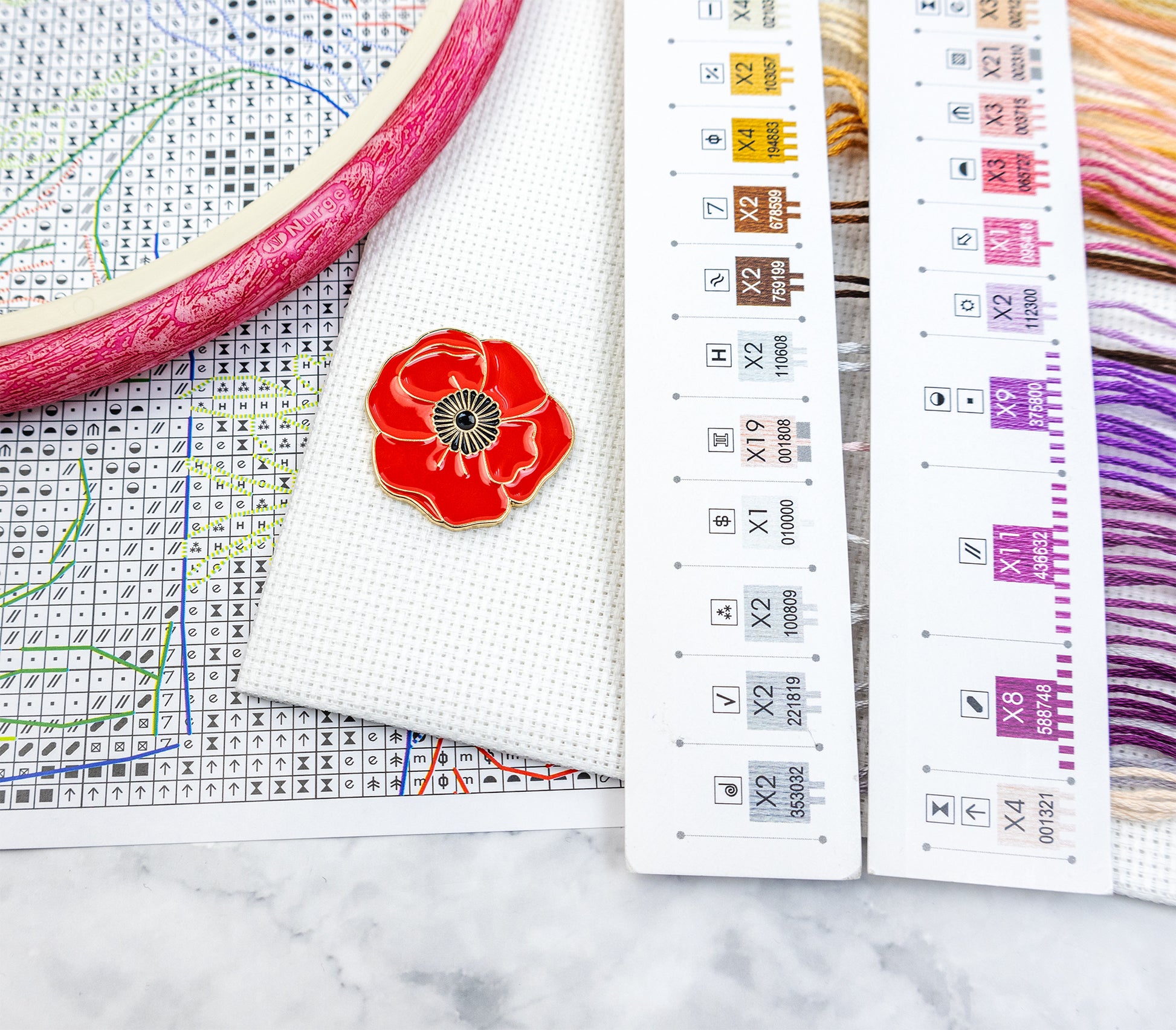 Embroidery hoop with red flower design, cross-stitch chart on a marble surface.