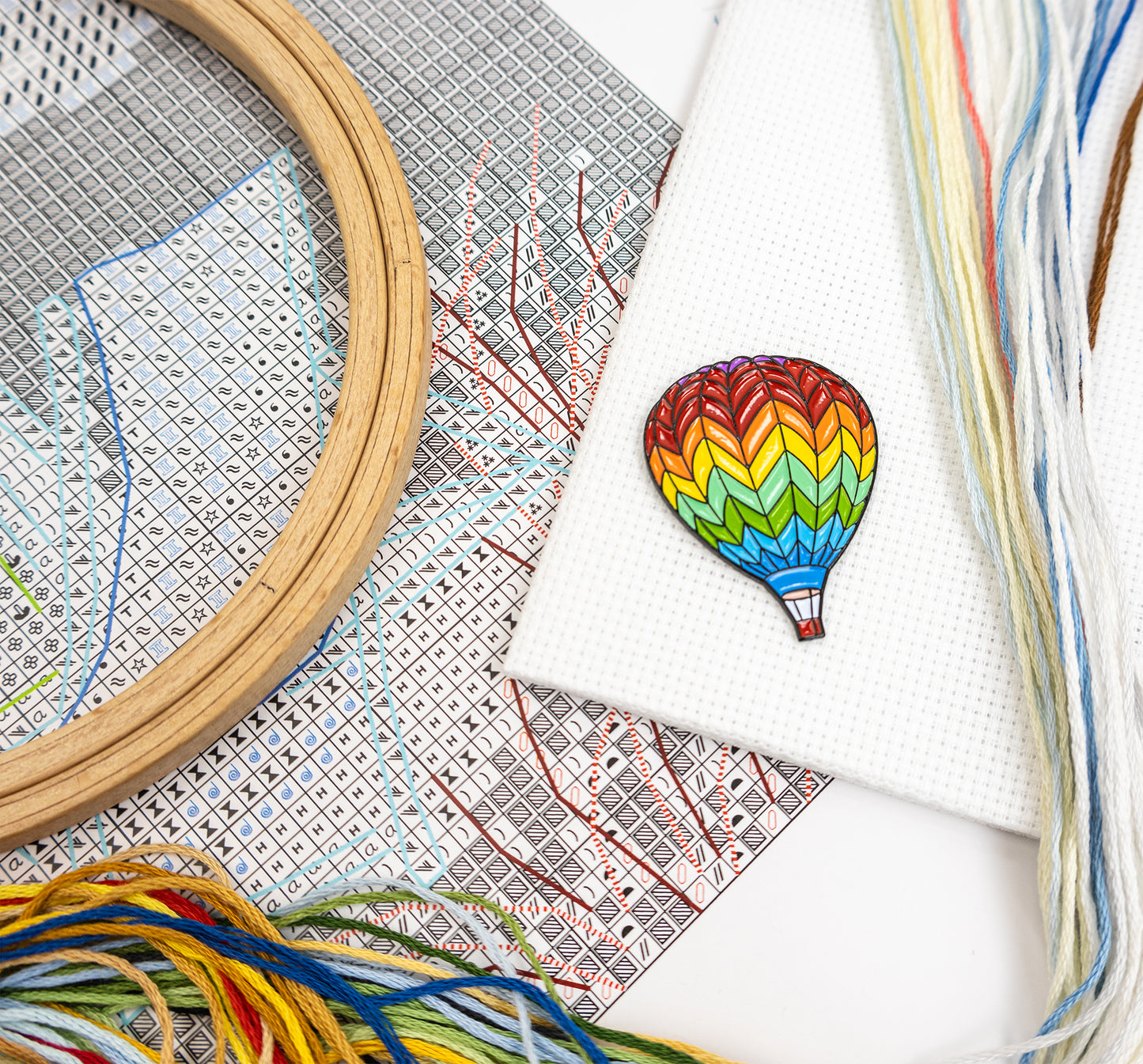 Embroidery hoop, cross-stitch pattern, and colorful threads with a completed embroidery of a rainbow hot air balloon.