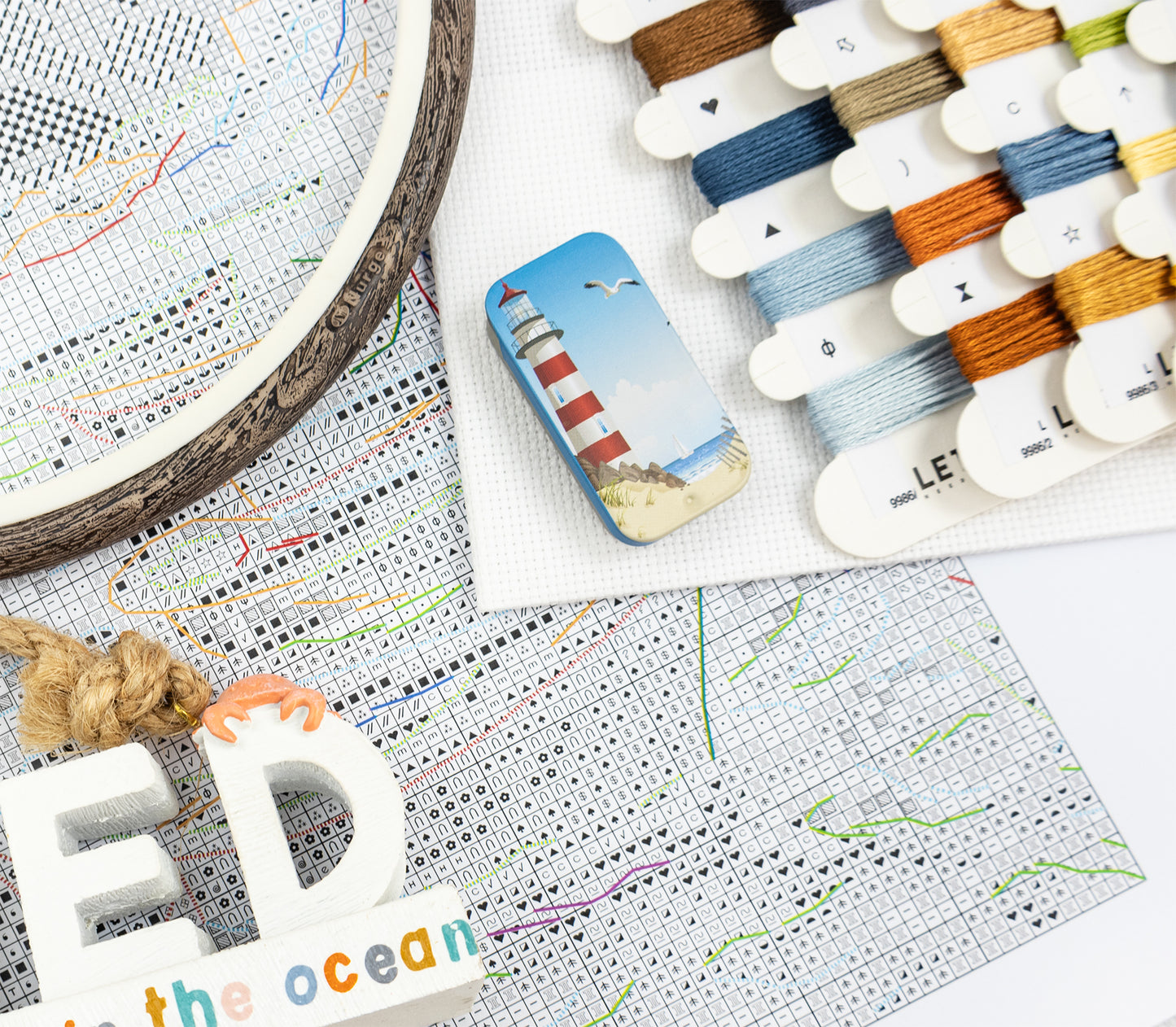 Embroidery hoop with cross-stitch pattern, thread spools, and a lighthouse-themed card on a map background.