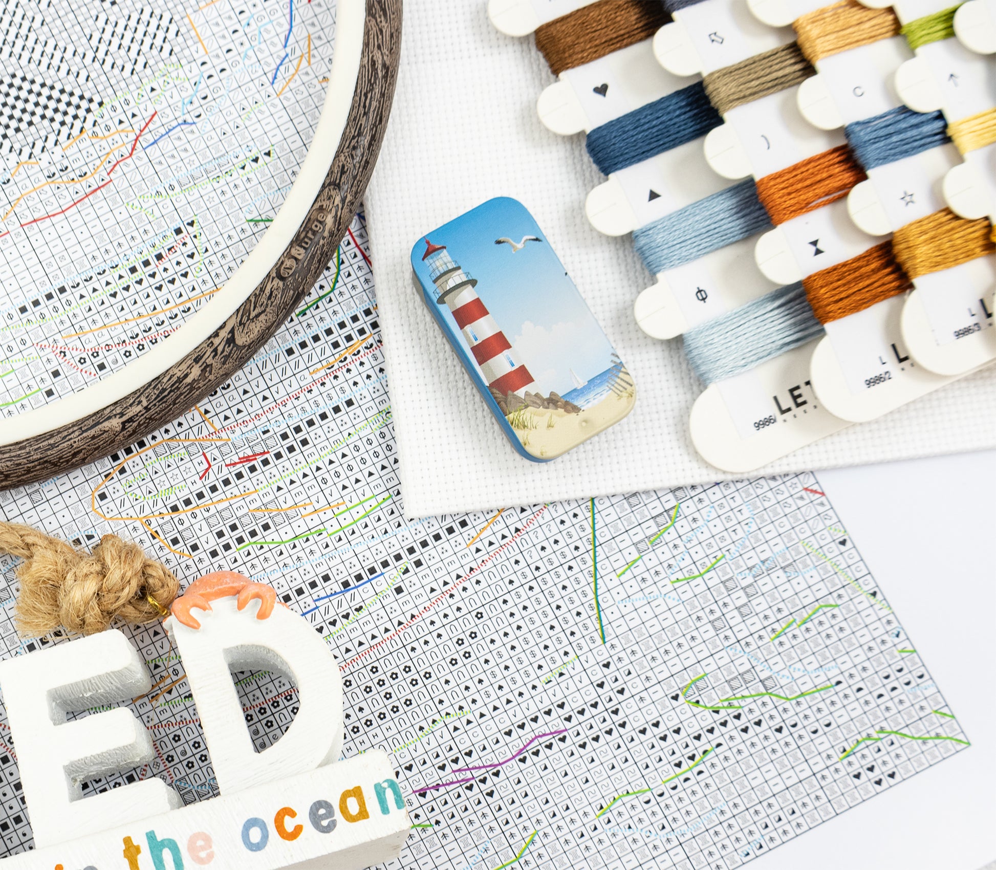 Embroidery hoop with cross-stitch pattern, thread spools, and a lighthouse-themed card on a map background.