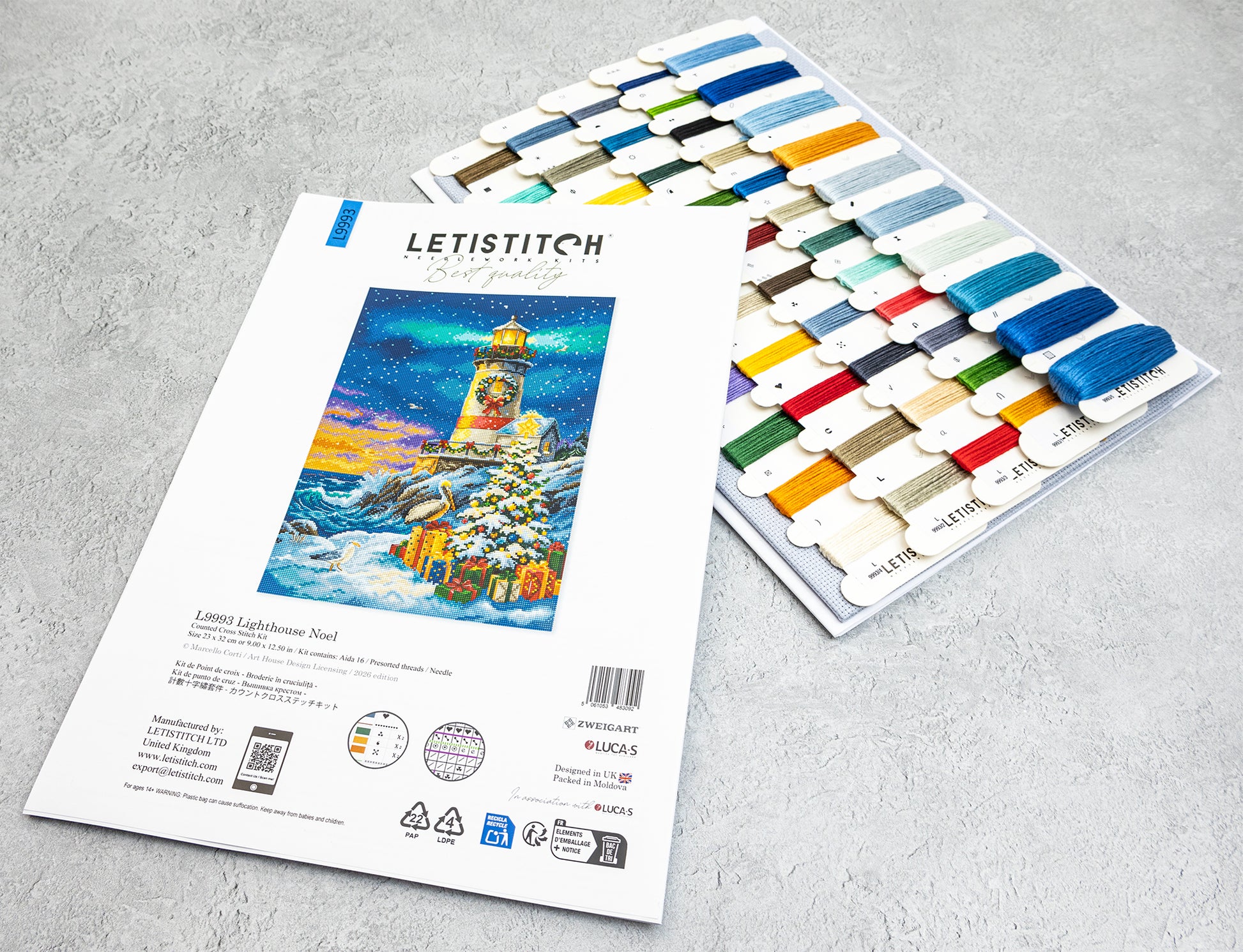 A Letistitch cross stitch kit for 'Lighthouse Noel' , displayed alongside a tray of colorful thread spools on a light gray textured background.