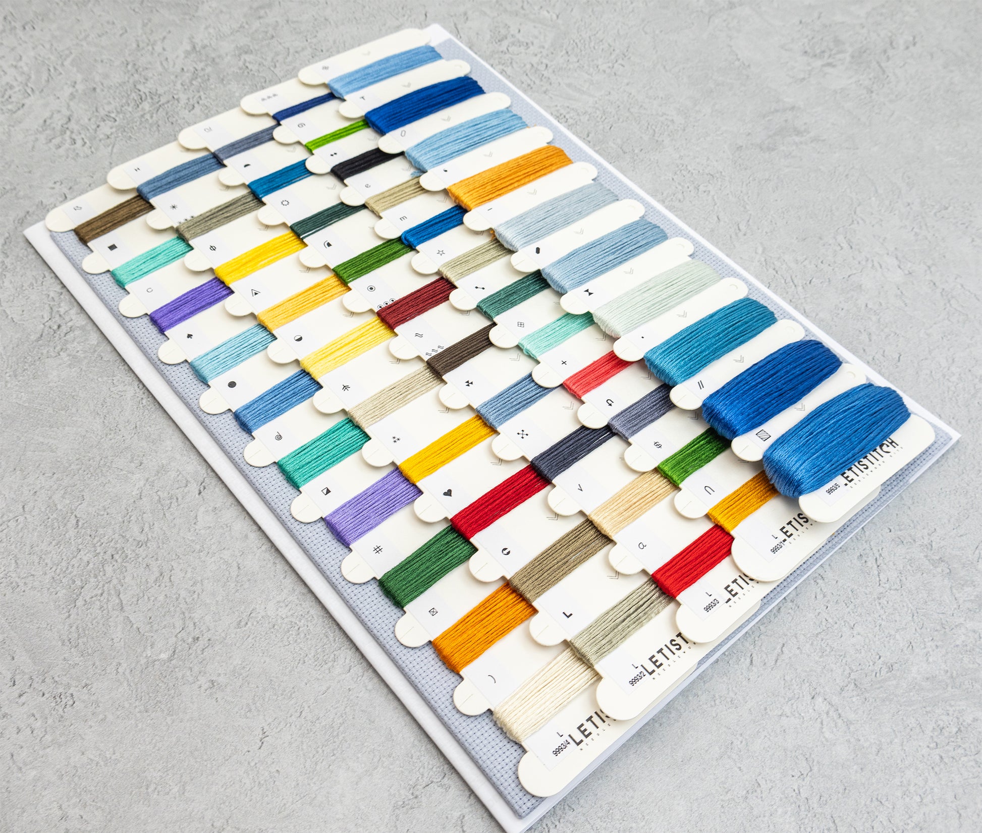 A display board filled with many small thread spools in assorted colors, photographed from above on a light gray textured surface. The spools are arranged in rows and include shades of blue, yellow, green, red, orange, purple, brown, black, white, and beige.