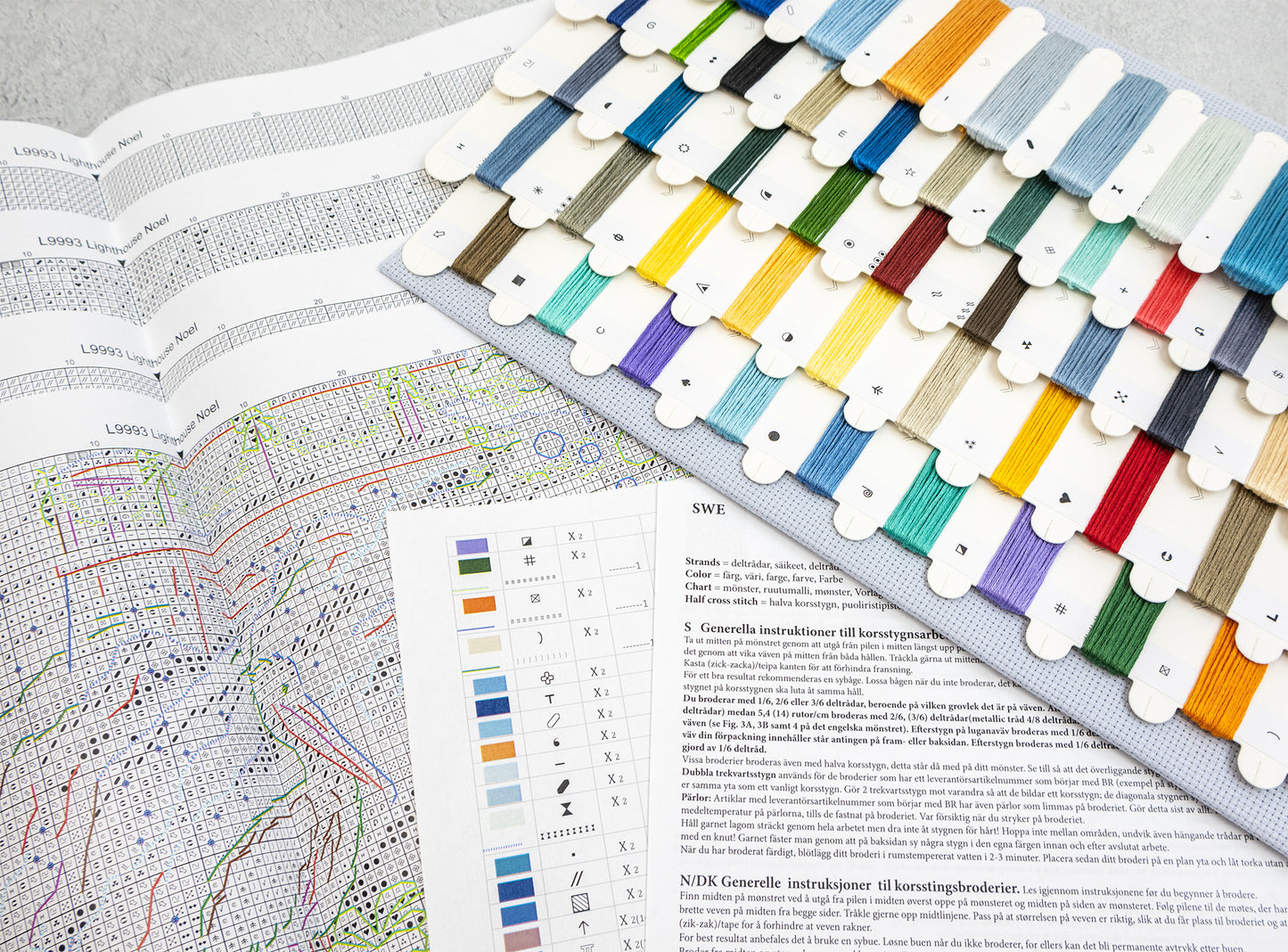 A fabric thread sampler card displaying dozens of colored embroidery floss threads arranged in rows, laid over an open embroidery pattern book with a color chart and instructions.