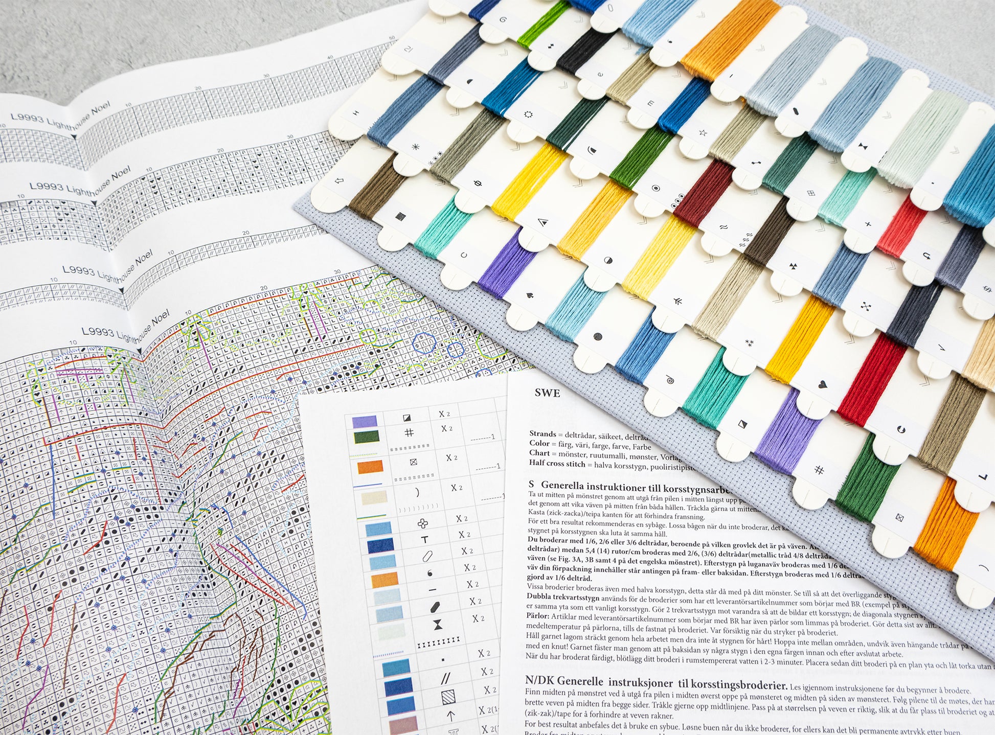 A fabric thread sampler card displaying dozens of colored embroidery floss threads arranged in rows, laid over an open embroidery pattern book with a color chart and instructions.