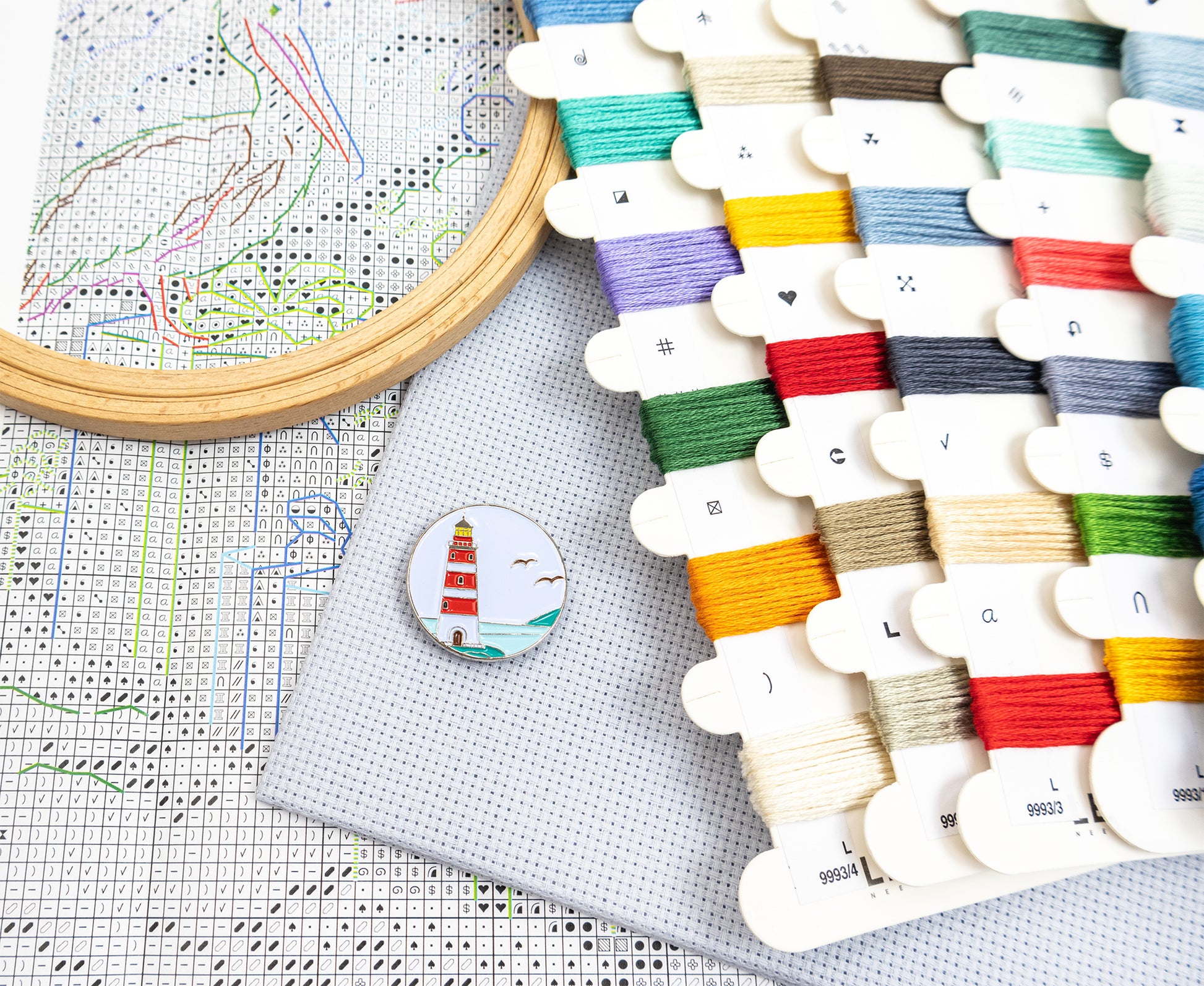 A styled flat-lay of embroidery supplies including a wooden hoop with thread, a printed cross-stitch pattern, a gray fabric square, and multiple white thread spools in assorted colors. A small round pin with a lighthouse design rests on the fabric.