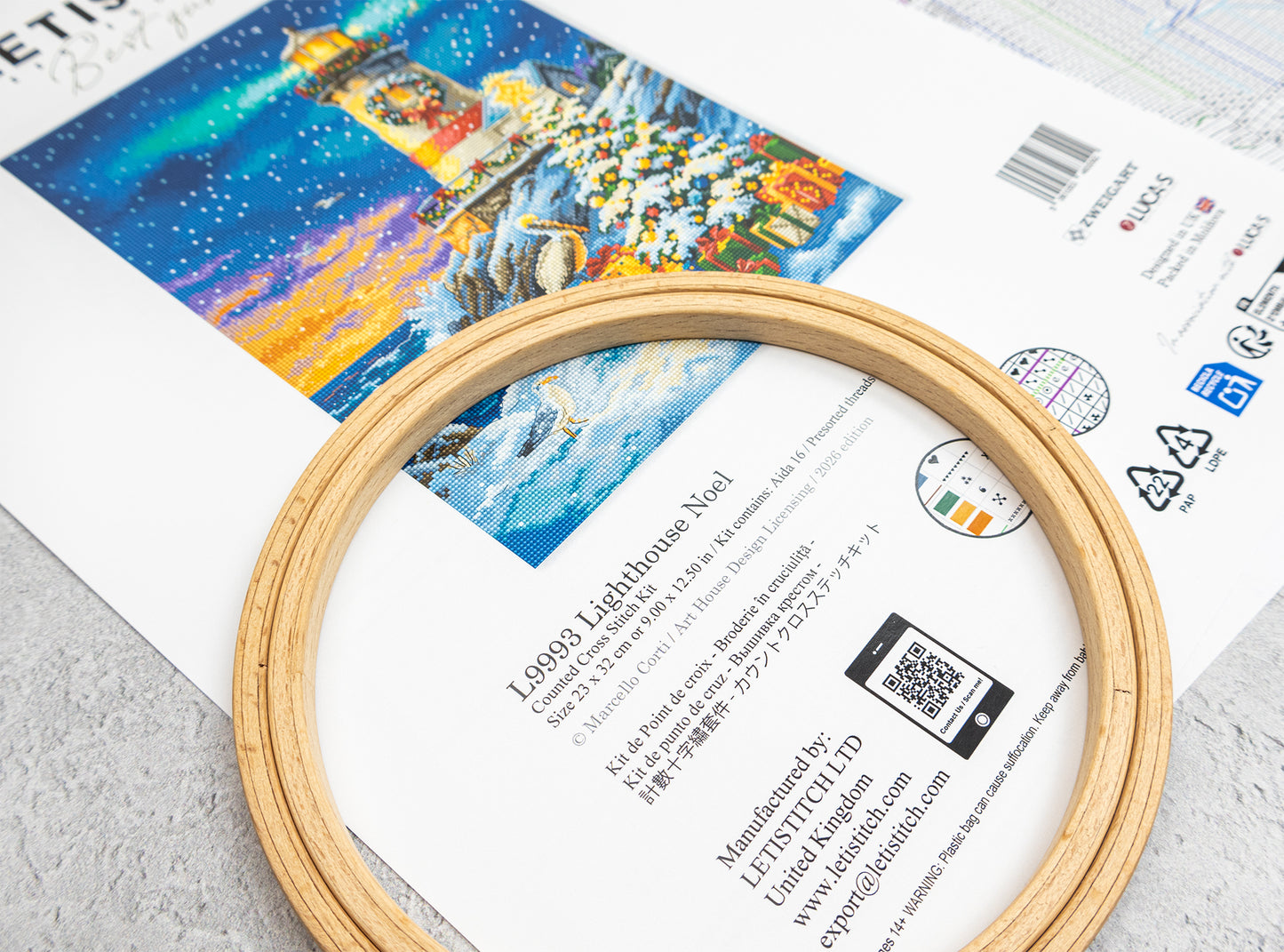 A wooden embroidery hoop placed over a printed pattern sheet featuring a colorful lighthouse design. The sheet includes product information, a barcode, and a QR code.