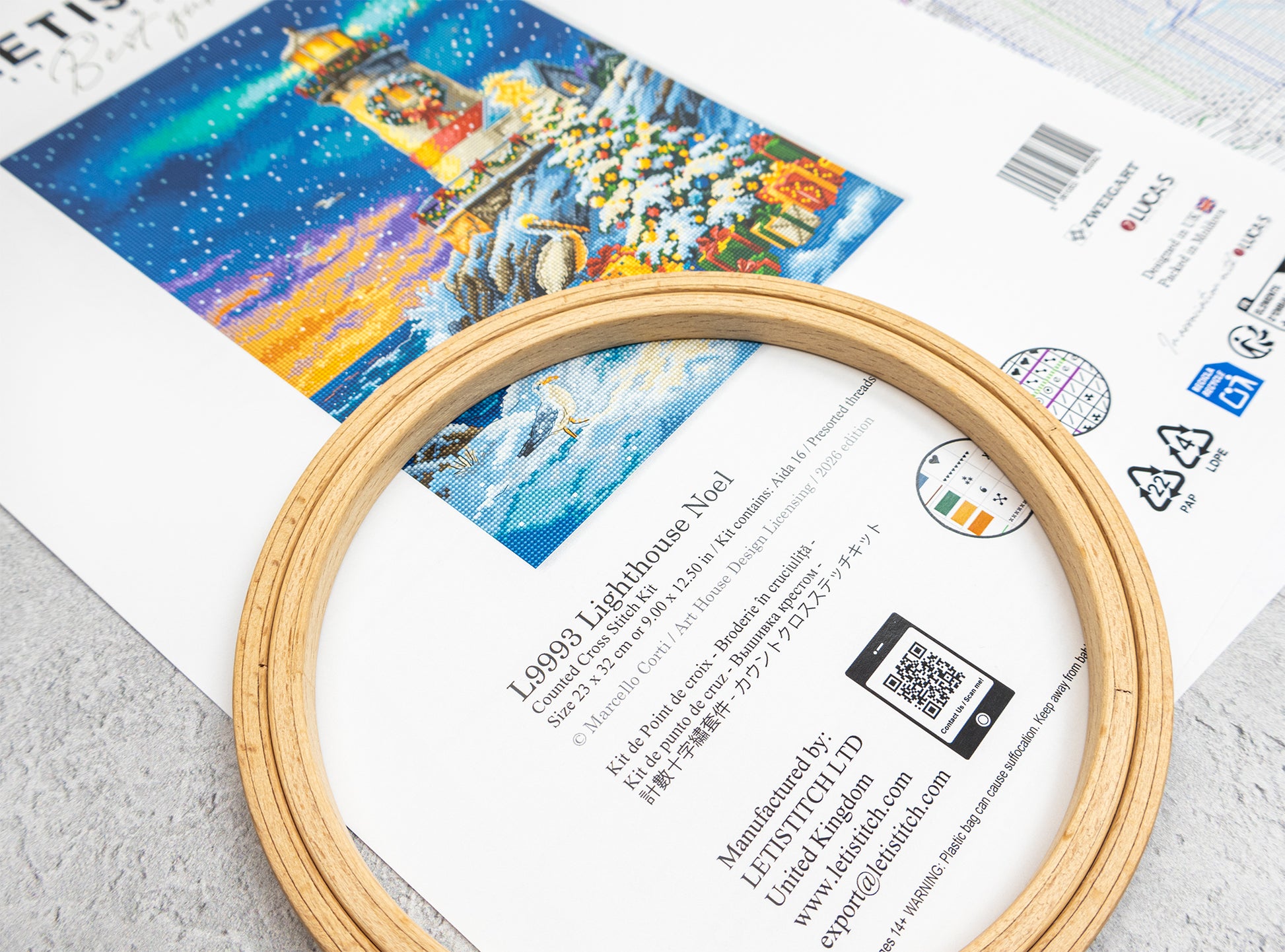 A wooden embroidery hoop placed over a printed pattern sheet featuring a colorful lighthouse design. The sheet includes product information, a barcode, and a QR code.