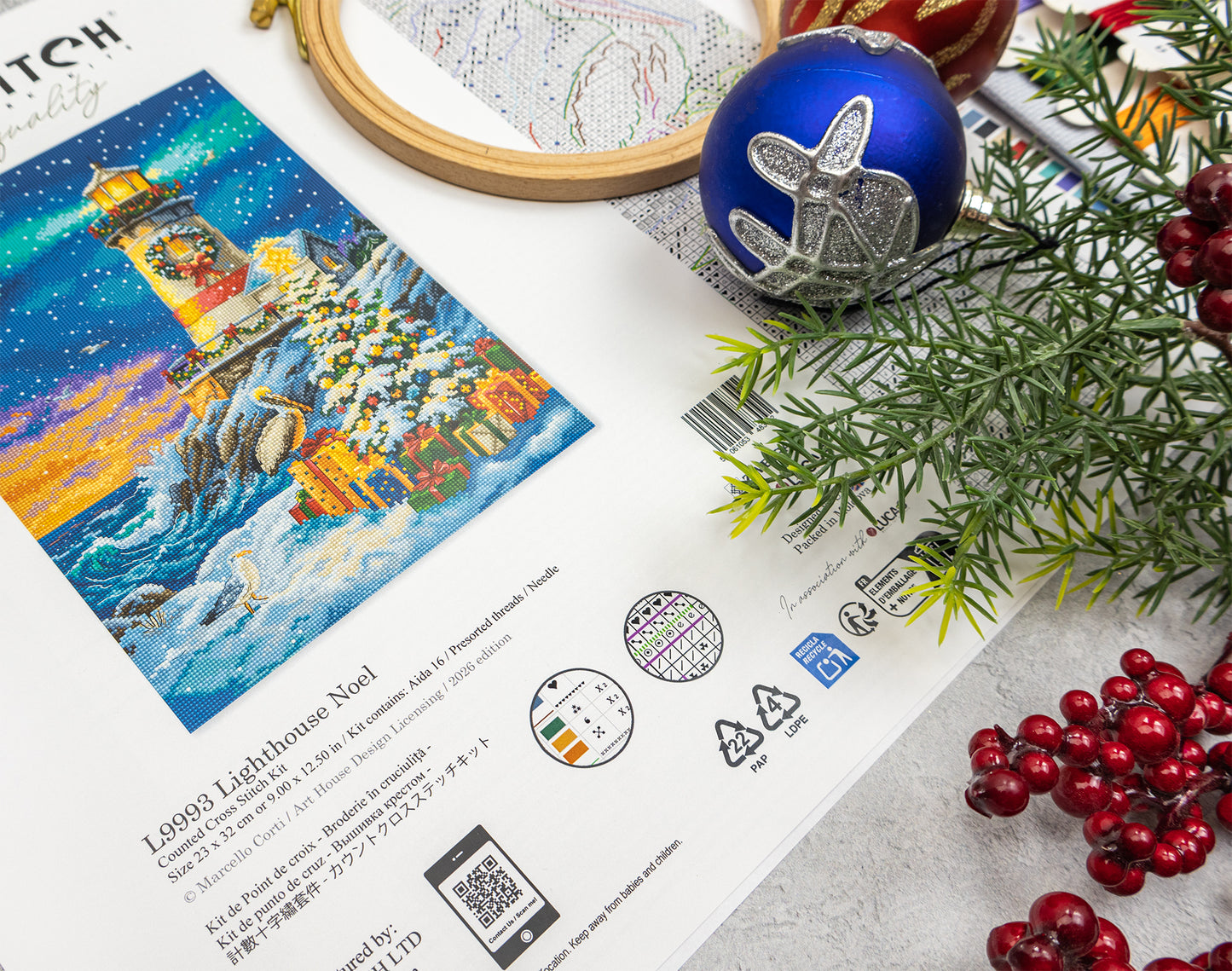 A printed craft kit box or art print featuring a colorful winter lighthouse illustration, styled with a blue ornament, silver ornament, green pine branches, and red berries on a light surface.