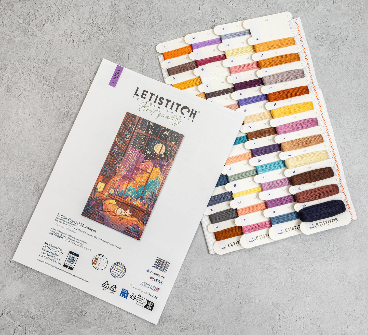 Letistitch Cross Stitch Kit package shown next to a thread sampler card displaying many thread colors, photographed on a light gray textured surface.