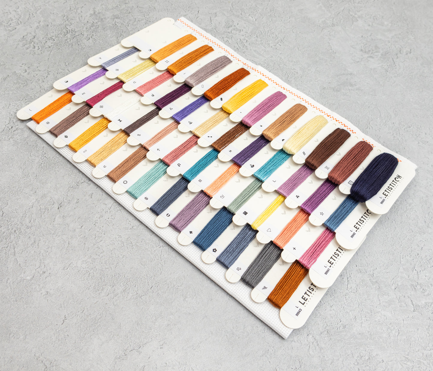 A display card filled with many small thread or embroidery floss swatches in assorted colors, photographed on a light gray textured surface. 