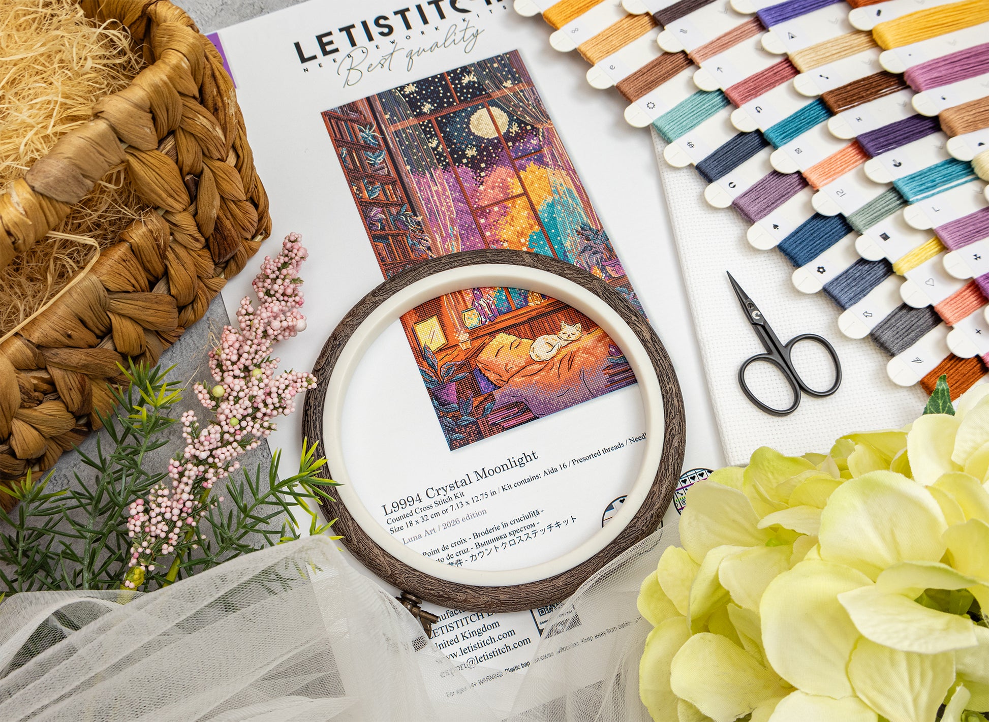 An embroidery pattern booklet and hoop displaying a colorful 'Crystal Moonlight' starry night design, styled in a flat lay with a basket, flowers, thread spools, and scissors.
