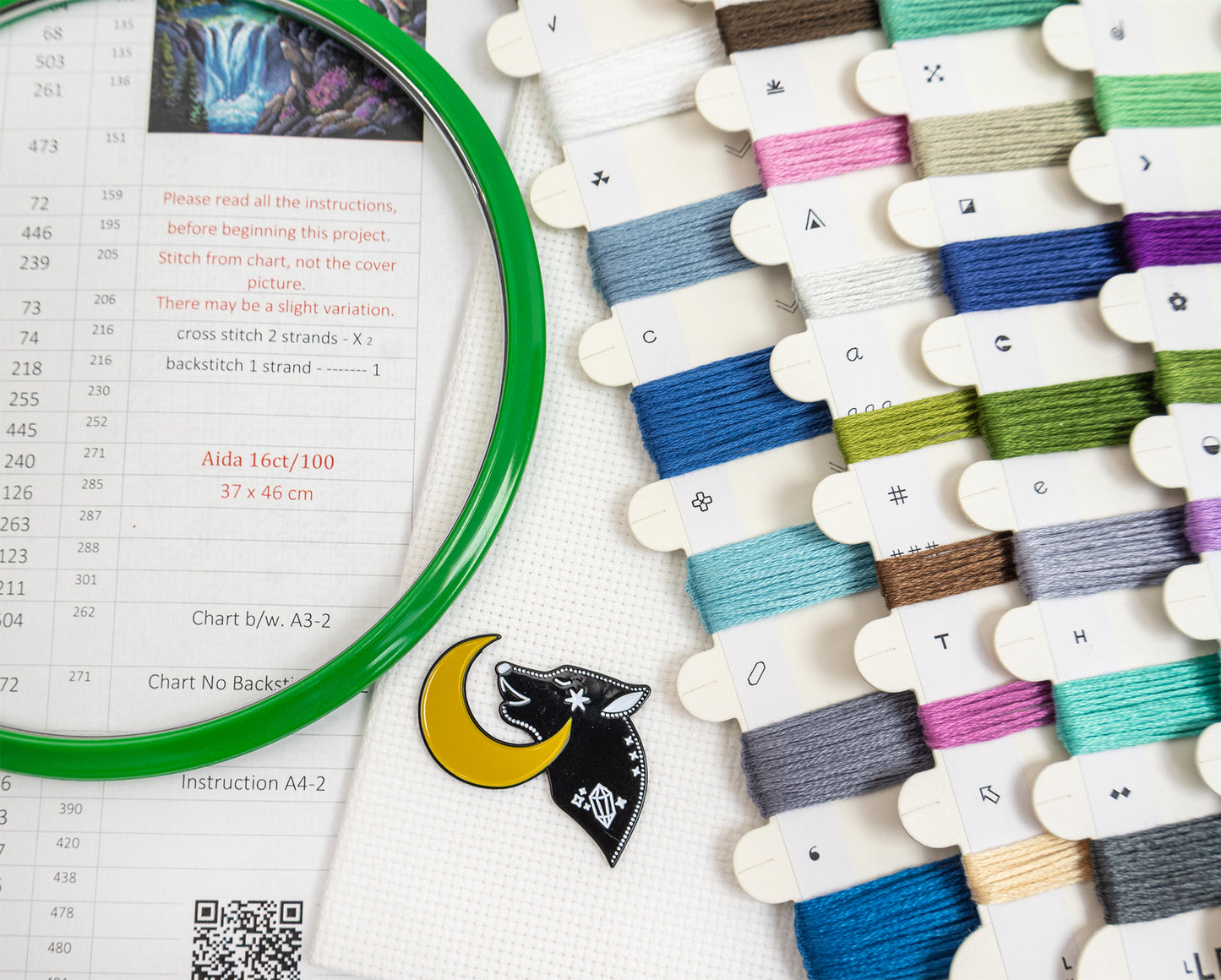 A close-up of a sewing or embroidery instruction sheet with a green circular magnifier, a small wolf emblem, and multiple thread spools in assorted colors arranged on a white surface.