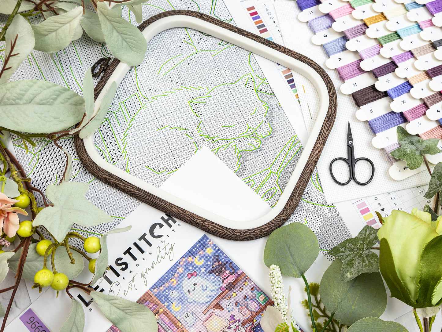 A styled flat-lay of embroidery supplies, featuring a square embroidery hoop with a white mesh cover and green stitched outline, surrounded by green leaves, small yellow berries, scissors, and a row of colorful thread spools on a white background.