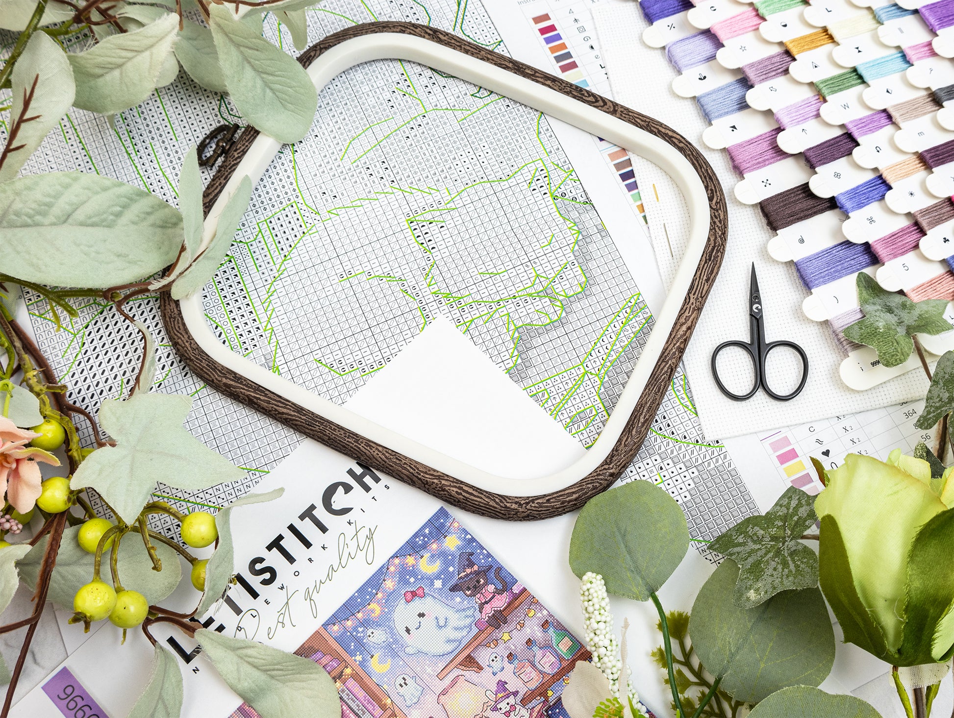 A styled flat-lay of embroidery supplies, featuring a square embroidery hoop with a white mesh cover and green stitched outline, surrounded by green leaves, small yellow berries, scissors, and a row of colorful thread spools on a white background.
