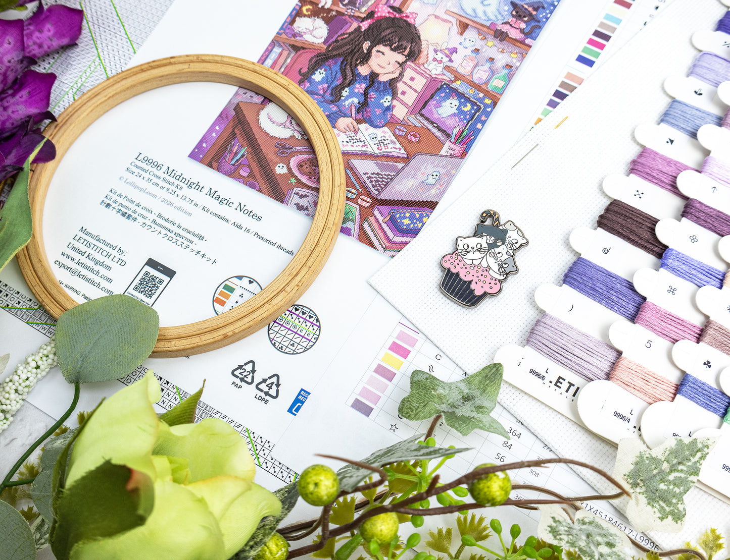 A styled flat-lay of embroidery thread swatches, a wooden hoop frame, and an illustrated pattern sheet featuring a girl in a shop. The scene includes decorative greenery and flowers, with the text 'Midnight Magic Notes' visible on the pattern insert.