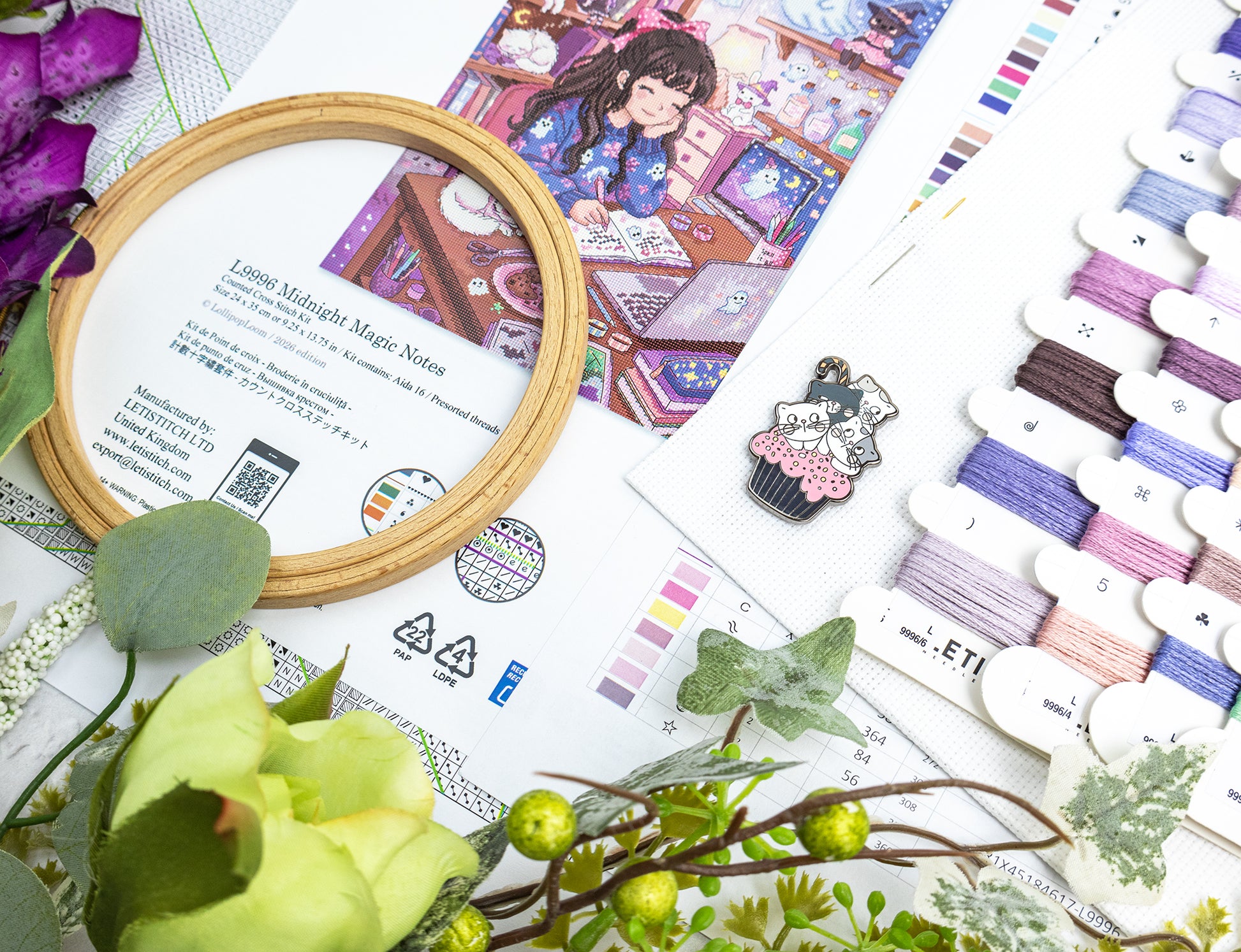 A styled flat-lay of embroidery thread swatches, a wooden hoop frame, and an illustrated pattern sheet featuring a girl in a shop. The scene includes decorative greenery and flowers, with the text 'Midnight Magic Notes' visible on the pattern insert.