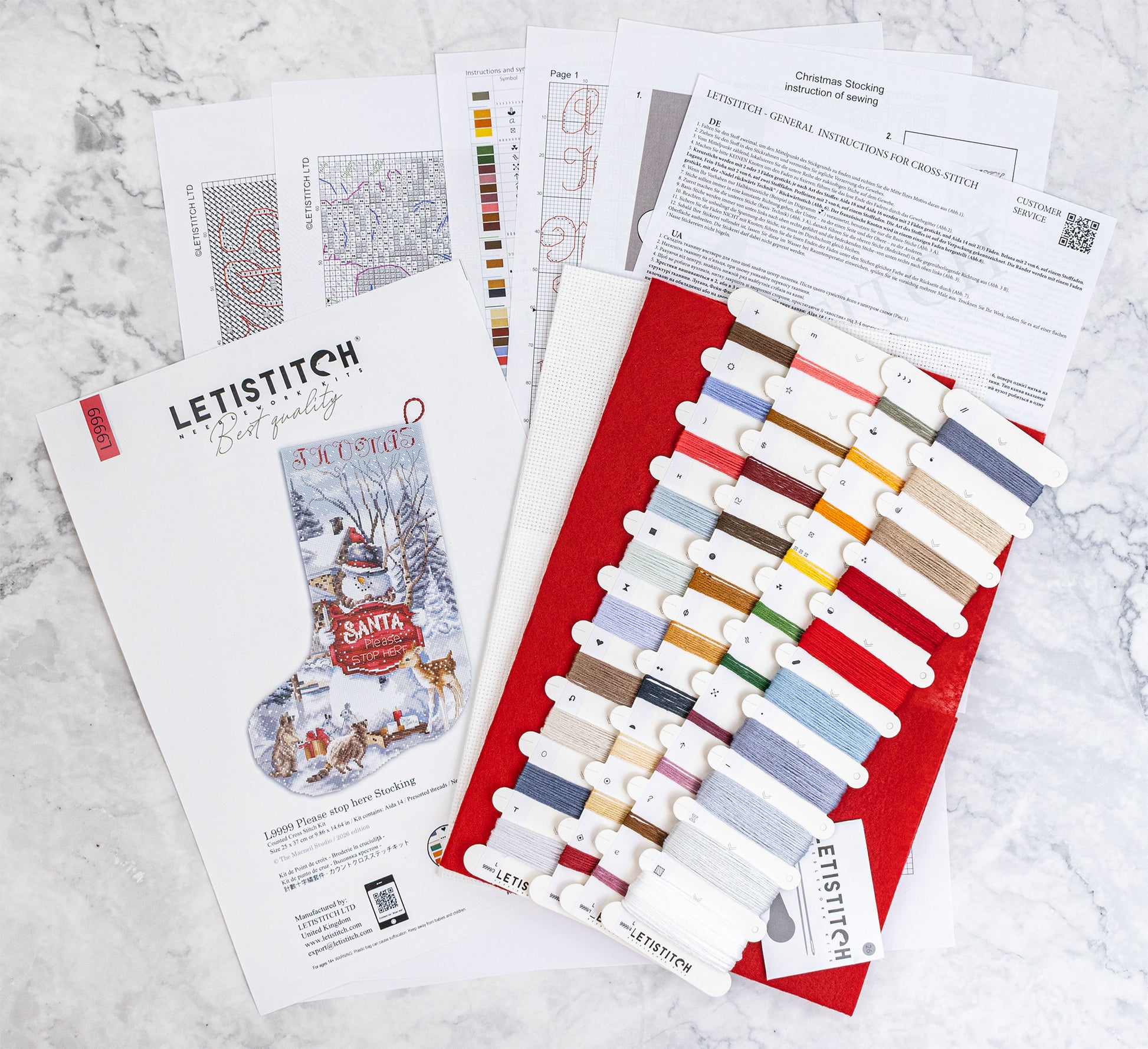 A flat lay of Letistitch Christmas Stocking embroidery kit components on a light marble surface, including the printed pattern booklet, instruction sheets, a red fabric backing, and multiple spools of thread in assorted colors.