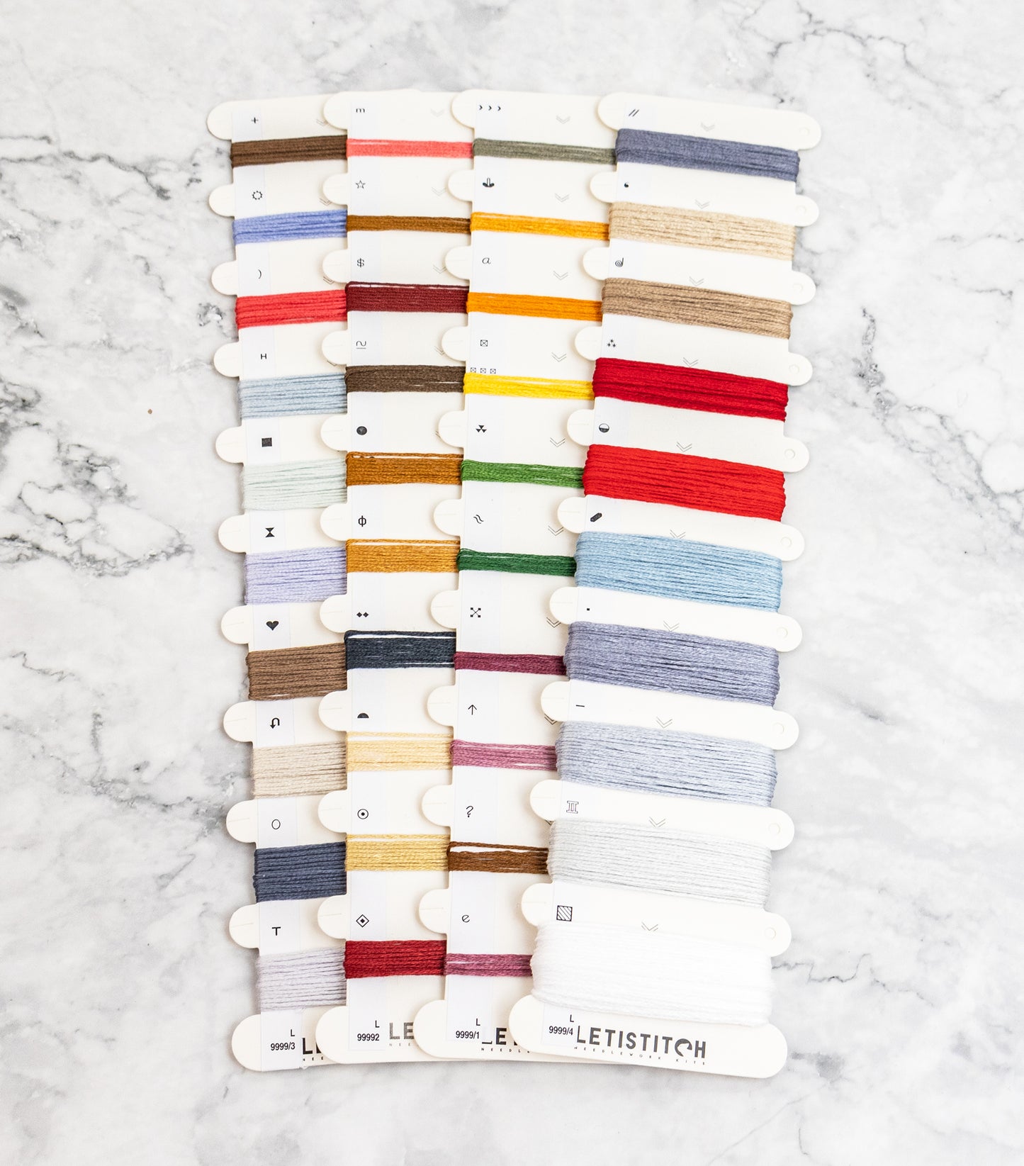 A set of embroidery floss spools arranged in three columns on a white marble background. The spools display a wide variety of colors, including red, blue, green, yellow, orange, brown, beige, and white.