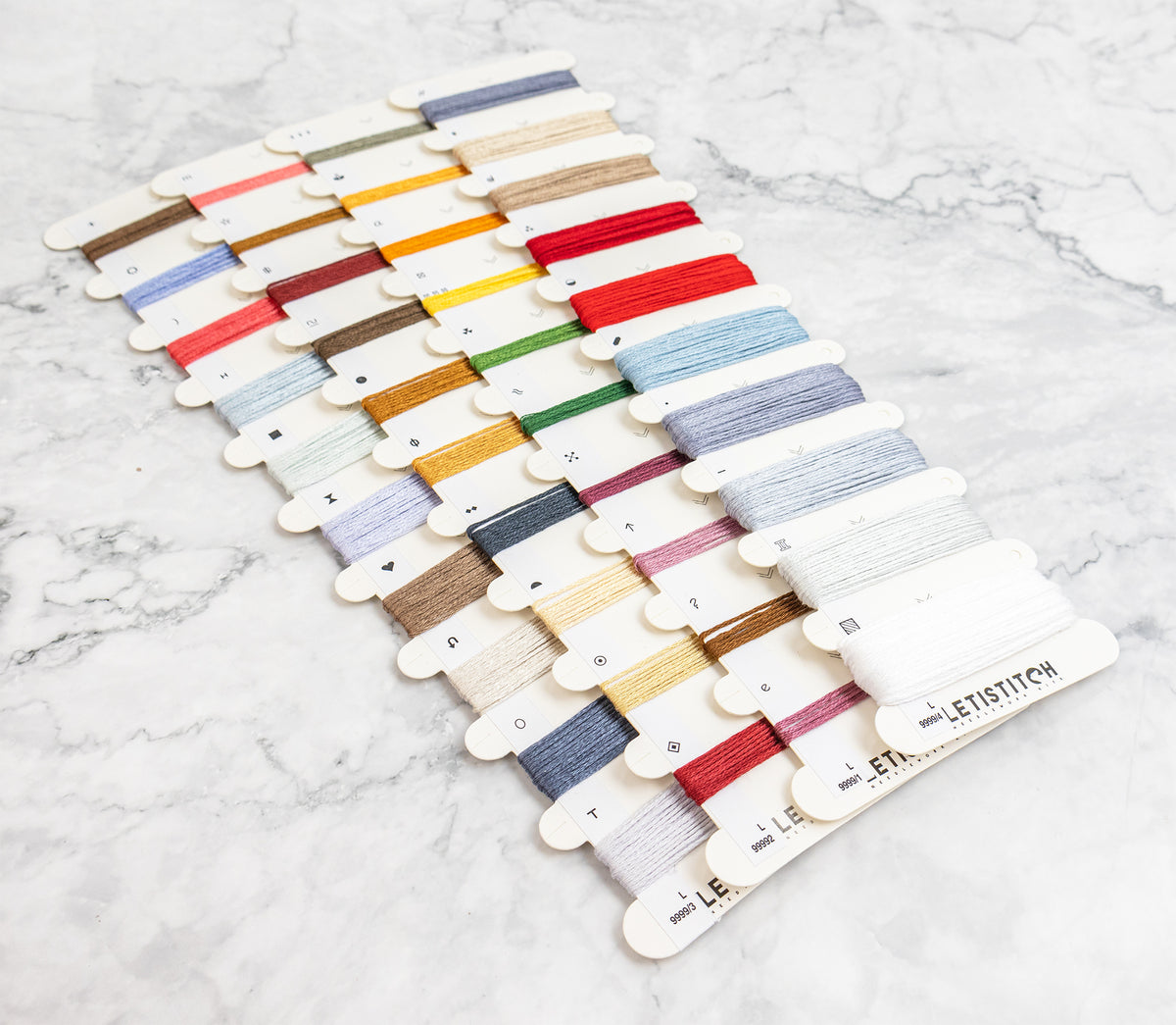 A set of white thread spool cards arranged in rows on a light marble surface, showing many thread colors including red, blue, green, yellow, gold, pink, brown, and white.