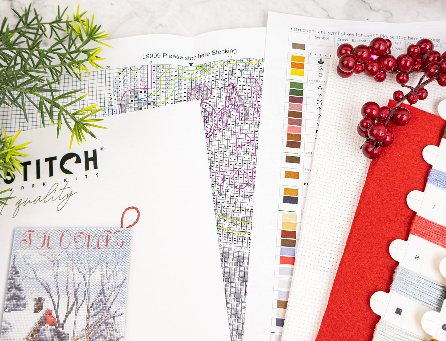 A flat lay of Christmas stocking kit materials on a light marble surface, including a printed pattern sheet, color chart, finished sample card, red fabric, white thread swatches, and holiday greenery and red berries.