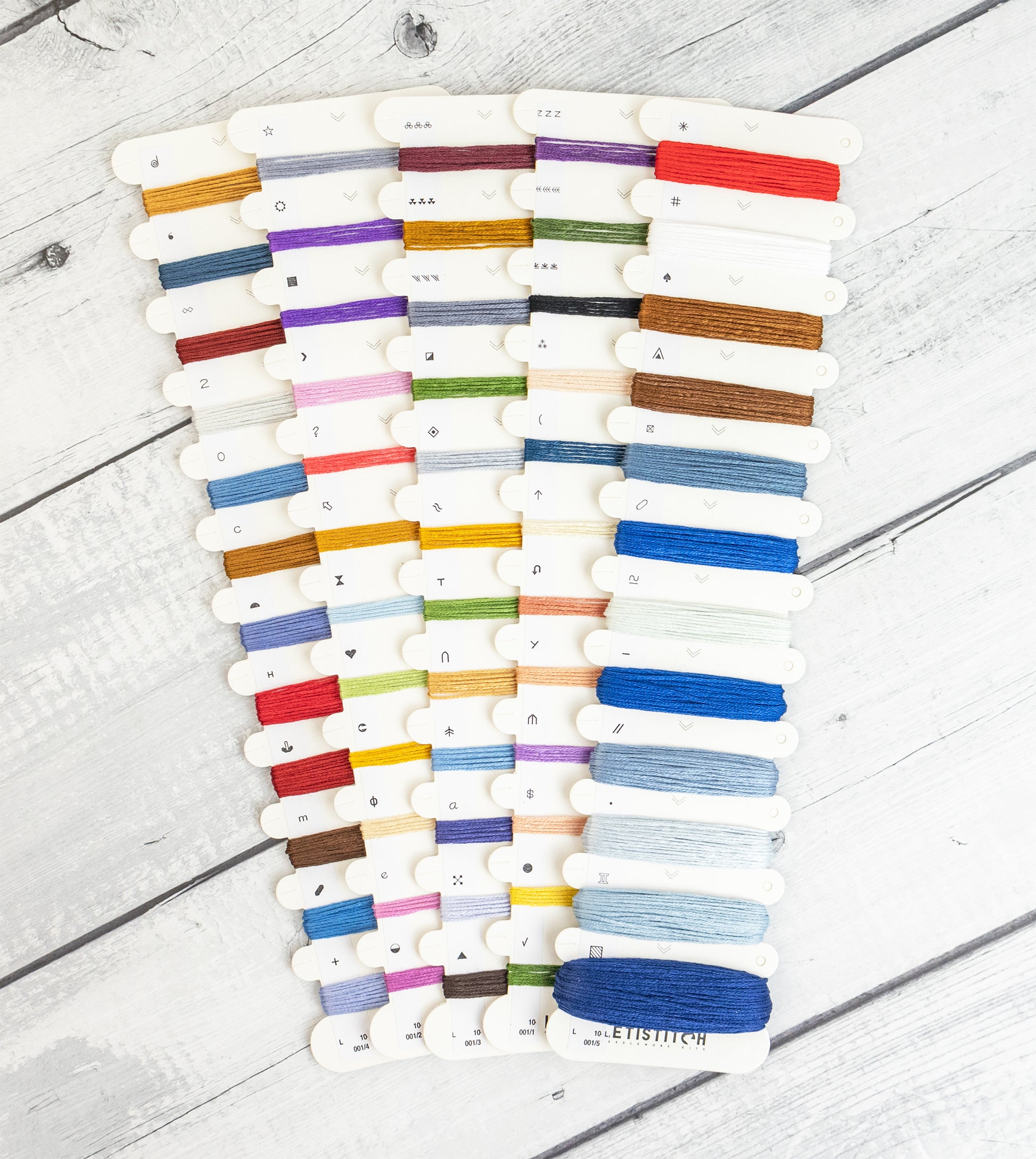 A set of white thread spools arranged in two columns on a white wood plank surface, displaying a wide variety of thread colors.
