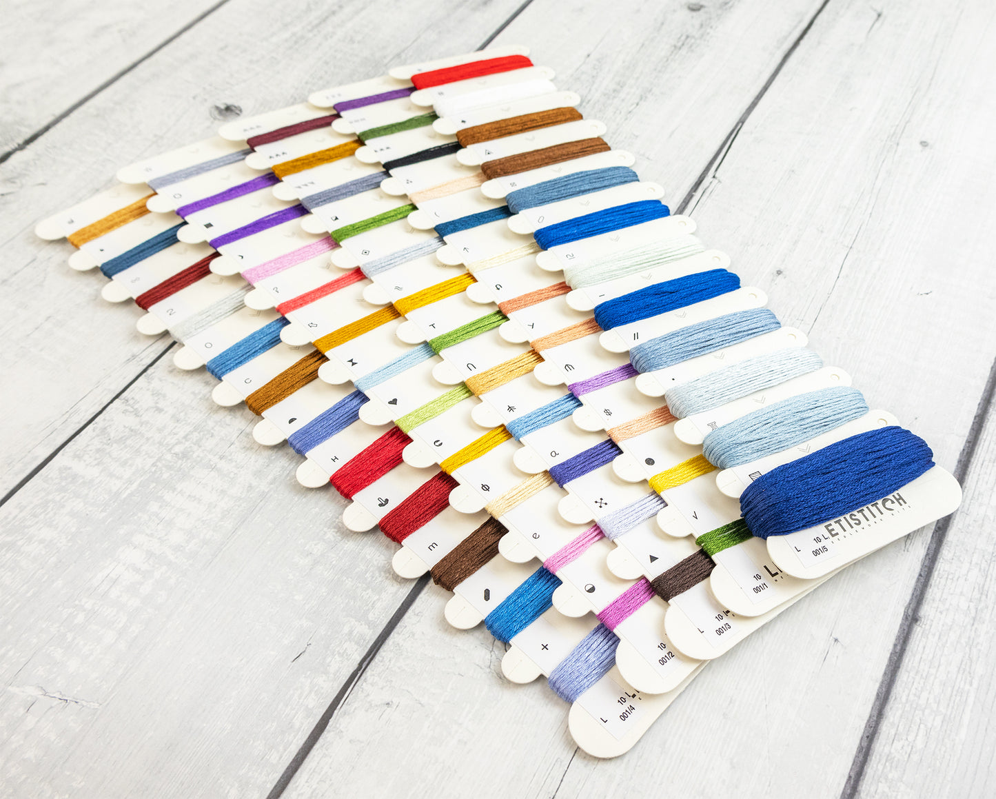A set of thread spools mounted on wooden sticks, arranged in rows on a light gray wood plank surface. The threads are in many different colors, including blue, red, yellow, green, purple, brown, and white, with a dark blue spool in the front right showing the brand name 'LETISTITCH'.
