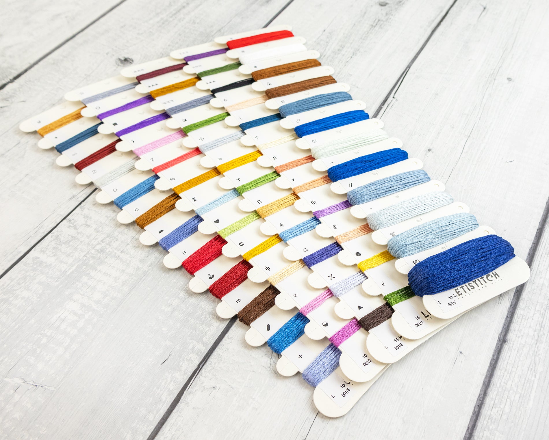 A set of thread spools mounted on wooden sticks, arranged in rows on a light gray wood plank surface. The threads are in many different colors, including blue, red, yellow, green, purple, brown, and white, with a dark blue spool in the front right showing the brand name 'LETISTITCH'.