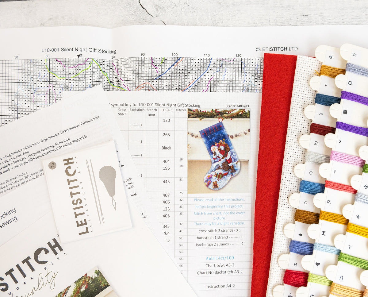A flat lay of cross-stitch kit components on a white surface, including printed pattern sheets, a symbol key card, a small booklet, a red fabric piece, a thread sampler card, and multiple spools of embroidery thread in assorted colors.