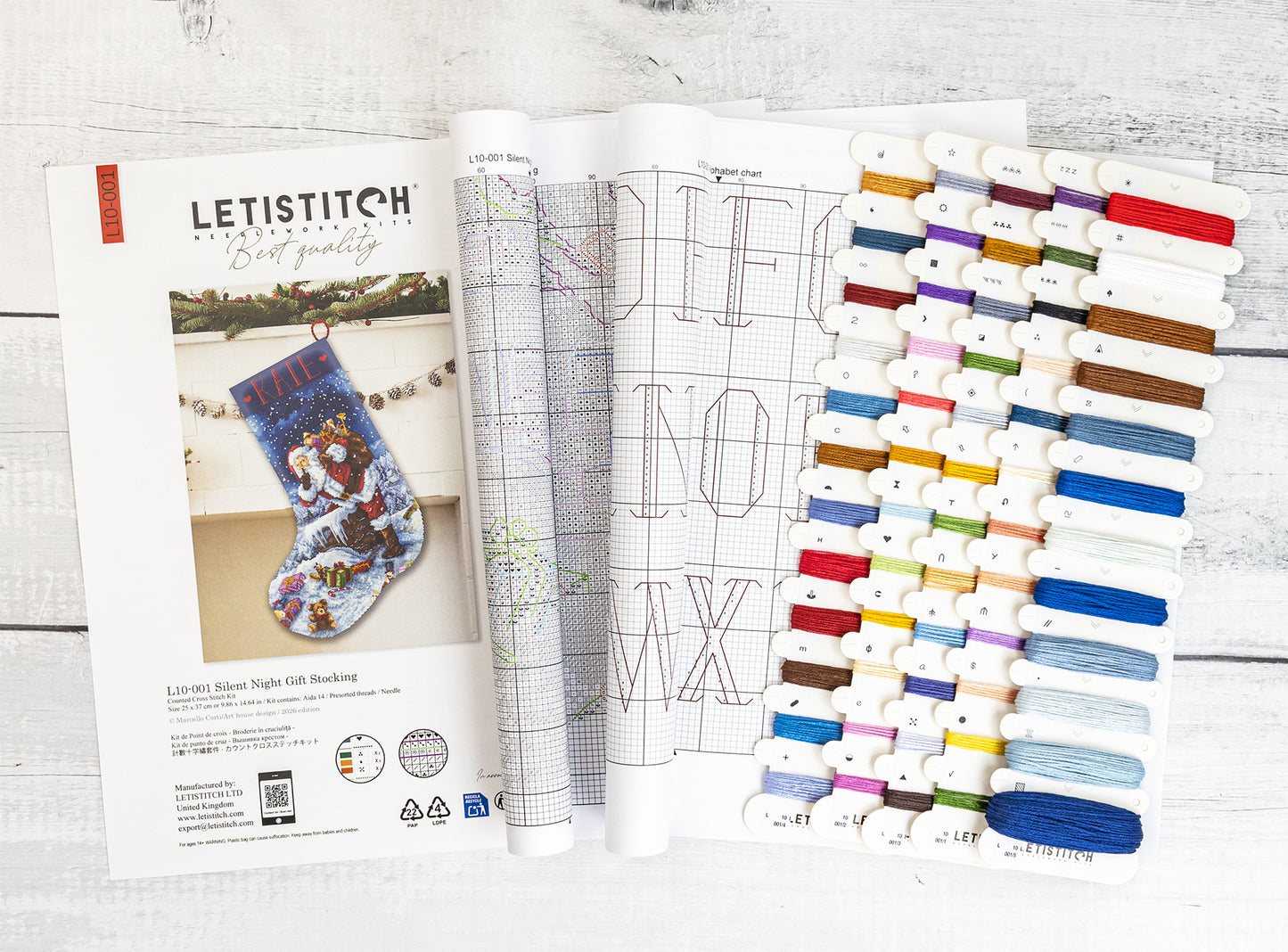 A Letistitch embroidery kit layout shown open on a white wood background, featuring a printed pattern booklet, a sample stitched stocking, and multiple thread color cards arranged in columns.