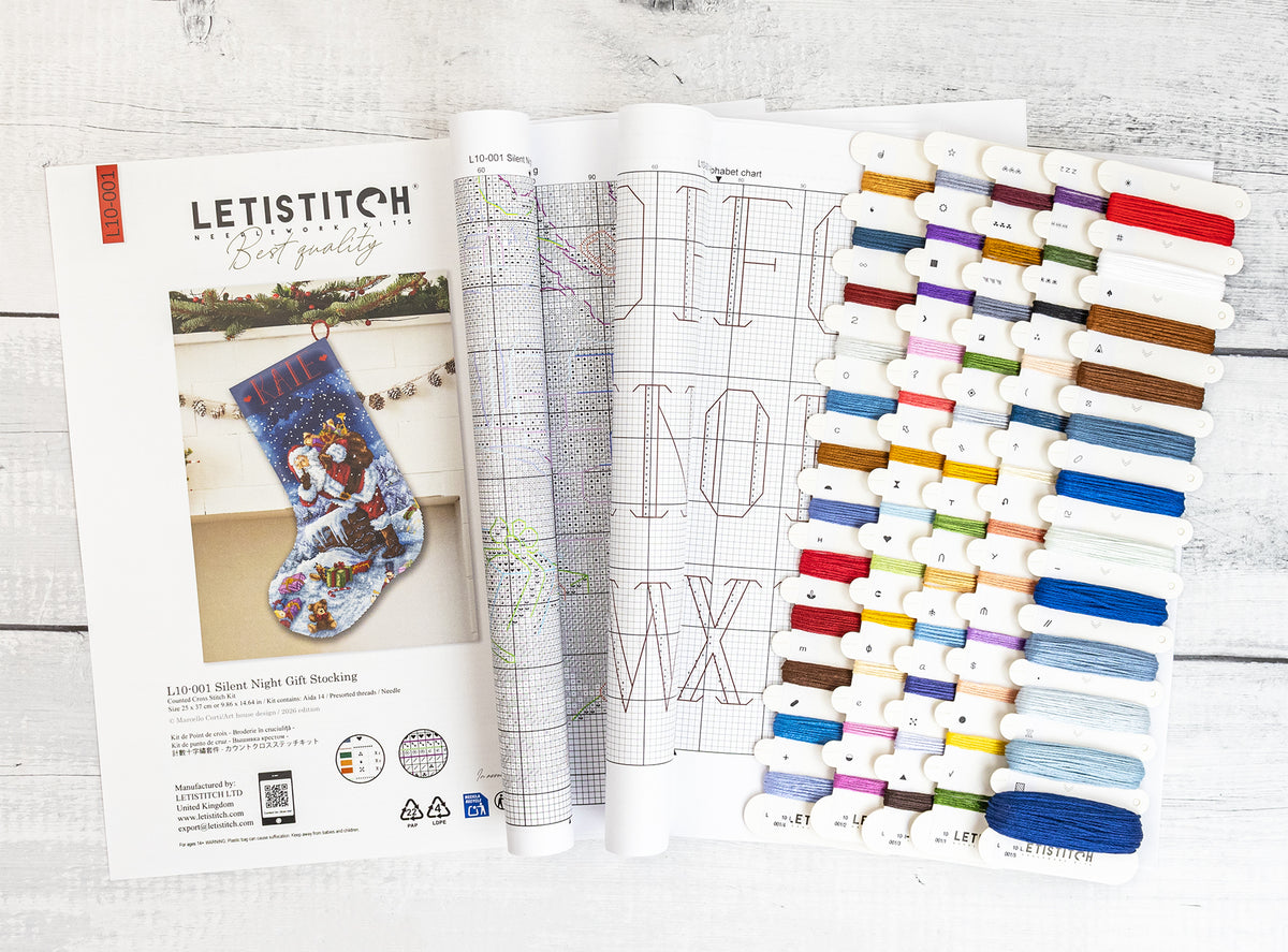 A Letistitch embroidery kit layout shown open on a white wood background, featuring a printed pattern booklet, a sample stitched stocking, and multiple thread color cards arranged in columns.