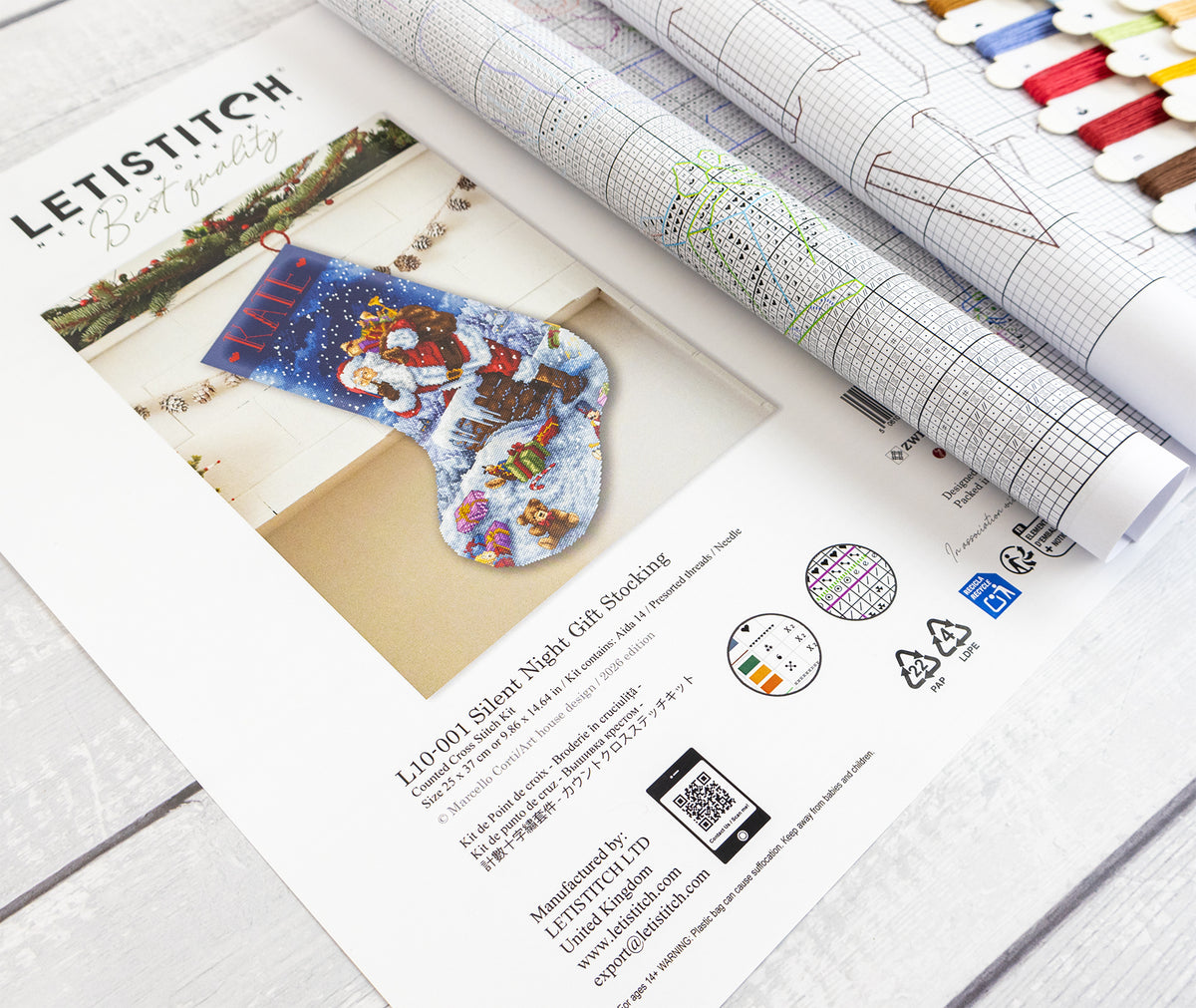 A printed Letistitch Christmas stocking kit booklet and rolled chart pages laid on a light wood surface, with thread spools visible along the top edge.