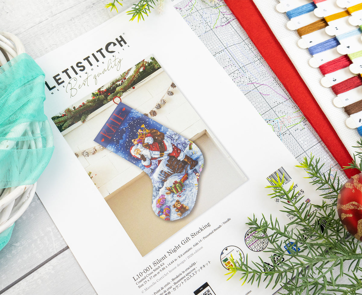 A printed sewing pattern booklet for a Letistitch Christmas stocking lies on a light wood surface, surrounded by craft props including spools of thread, a red ribbon, green pine branches, and a small ornament. The cover shows a completed blue stocking with festive embroidery.