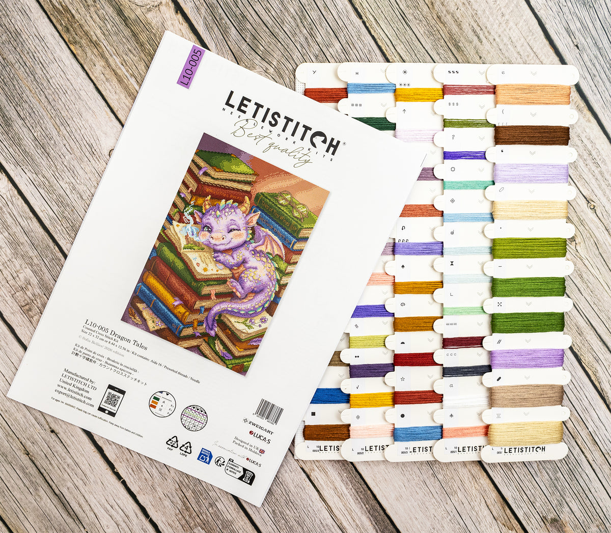A flat-lay of the LETISTITCH Embroidery Kit showing the white instruction booklet with a colorful dragon illustration, positioned next to a large array of colorful thread spools on a rustic wooden background.