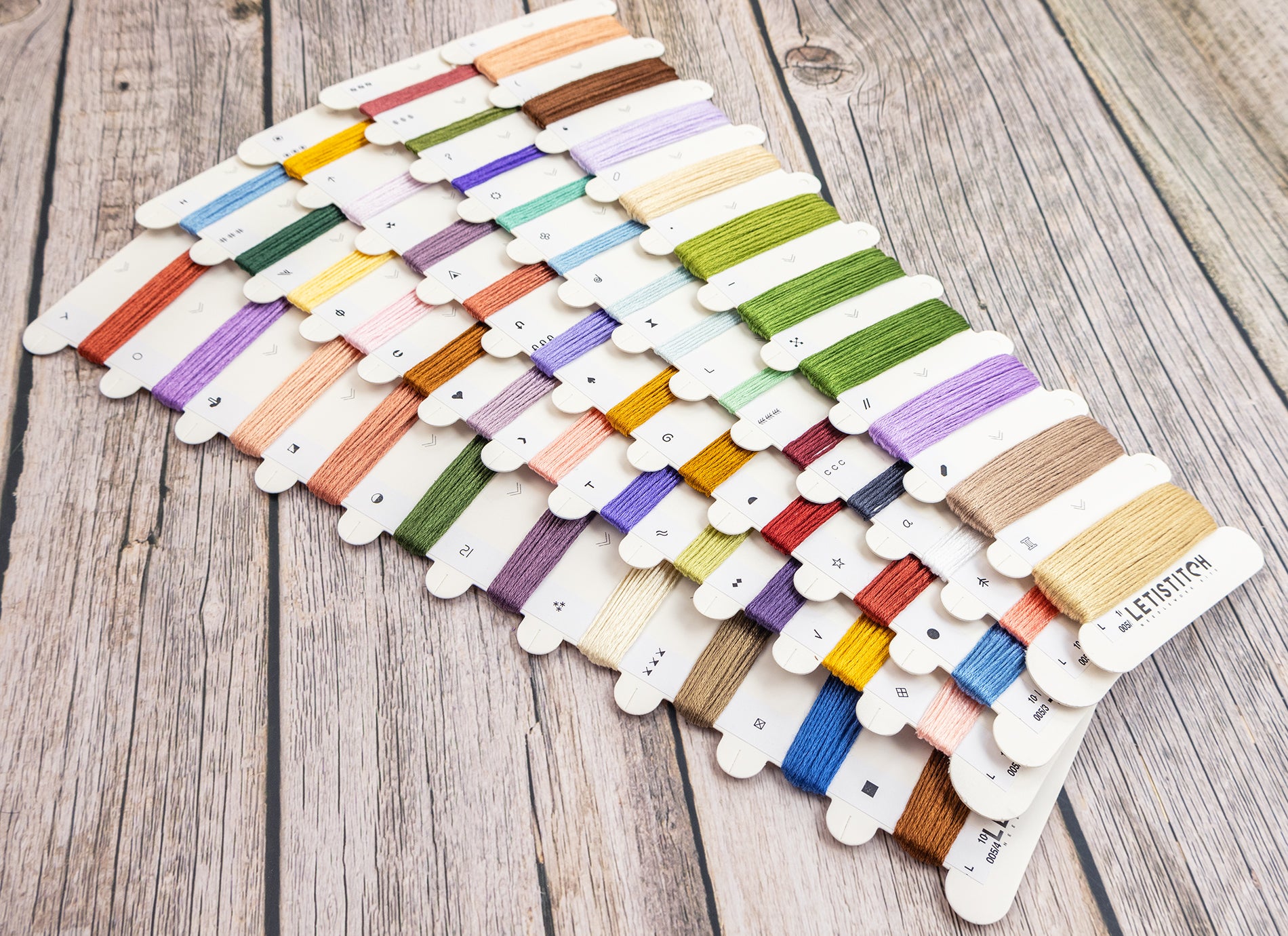 A set of embroidery floss thread spools arranged in rows on a rustic wooden surface. The threads come in a wide variety of colors, including greens, blues, purples, pinks, yellows, and browns, each mounted on a small white card.