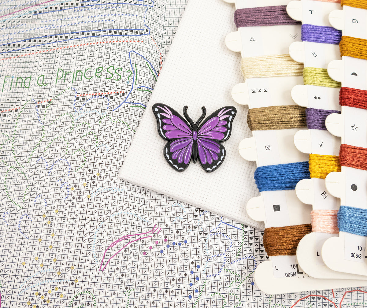 A purple and black butterfly-shaped enamel pin displayed on a white fabric swatch, next to a completed cross-stitch pattern sheet and multiple spools of embroidery thread in assorted colors.